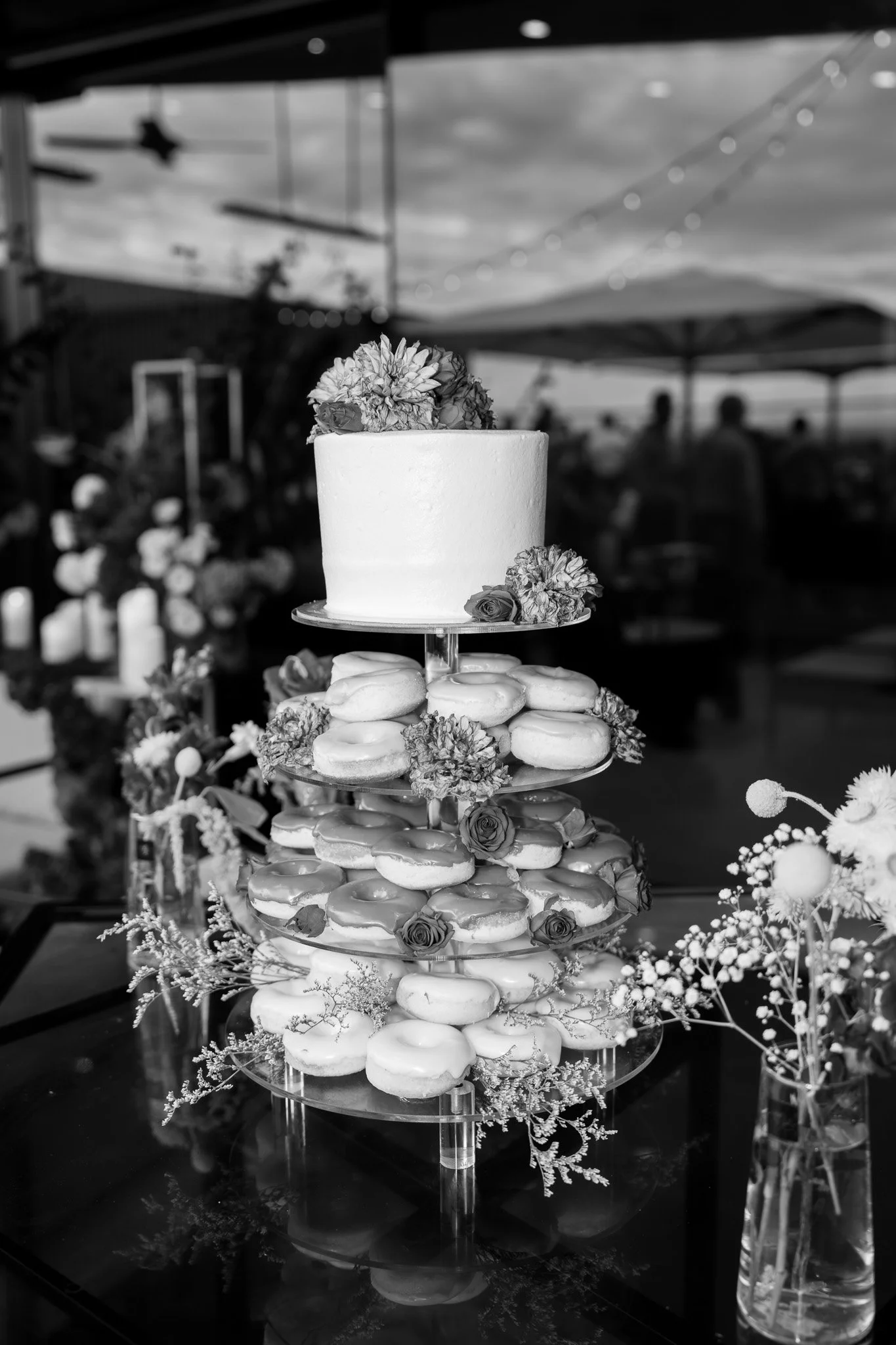 A tiered display with donuts and a cake decorated with flowers at an outdoor event.