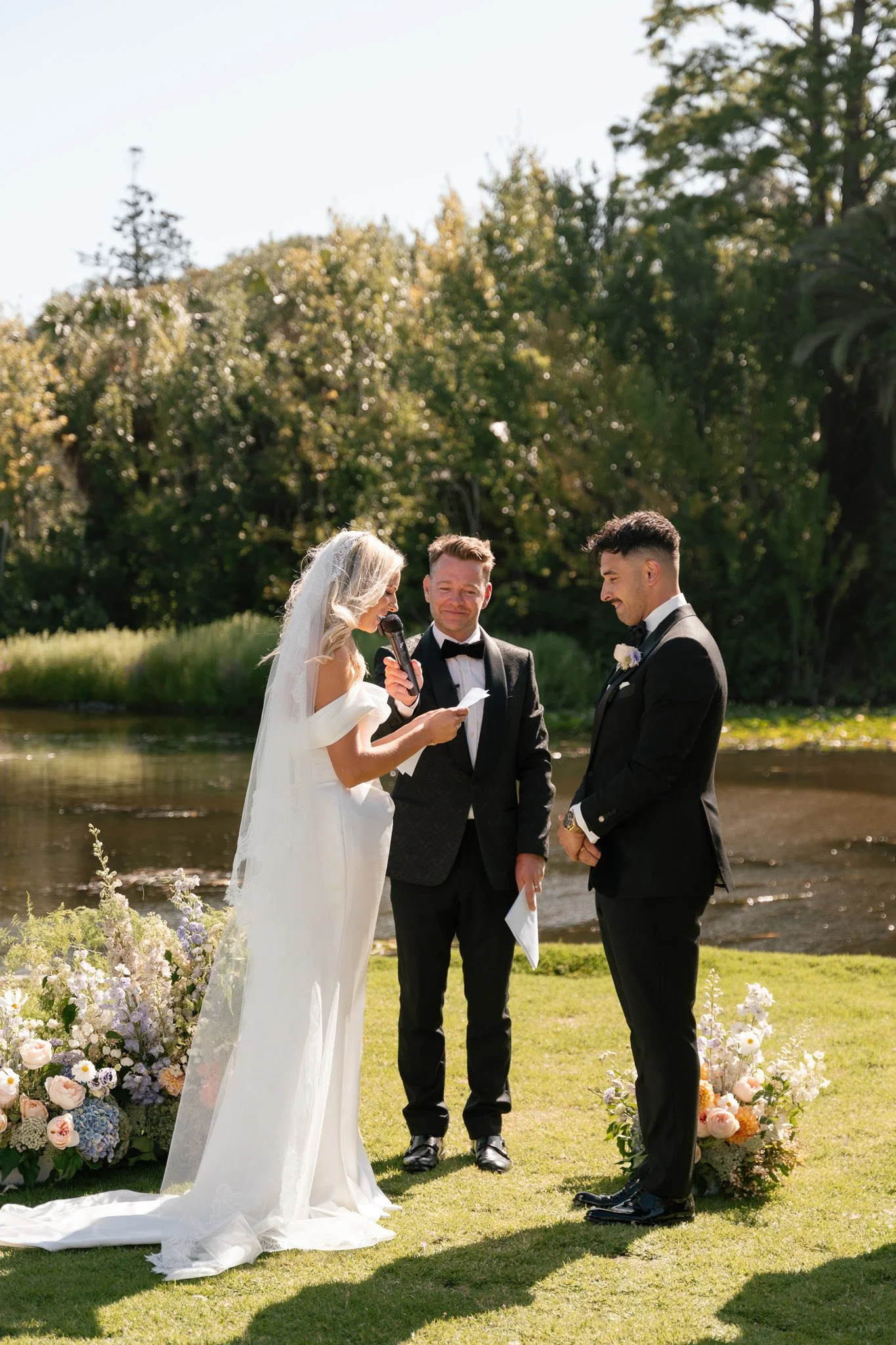 A bride and groom exchanging vows during an outdoor wedding ceremony near a lake, with officiant reading from a paper, surrounded by floral arrangements and green trees.