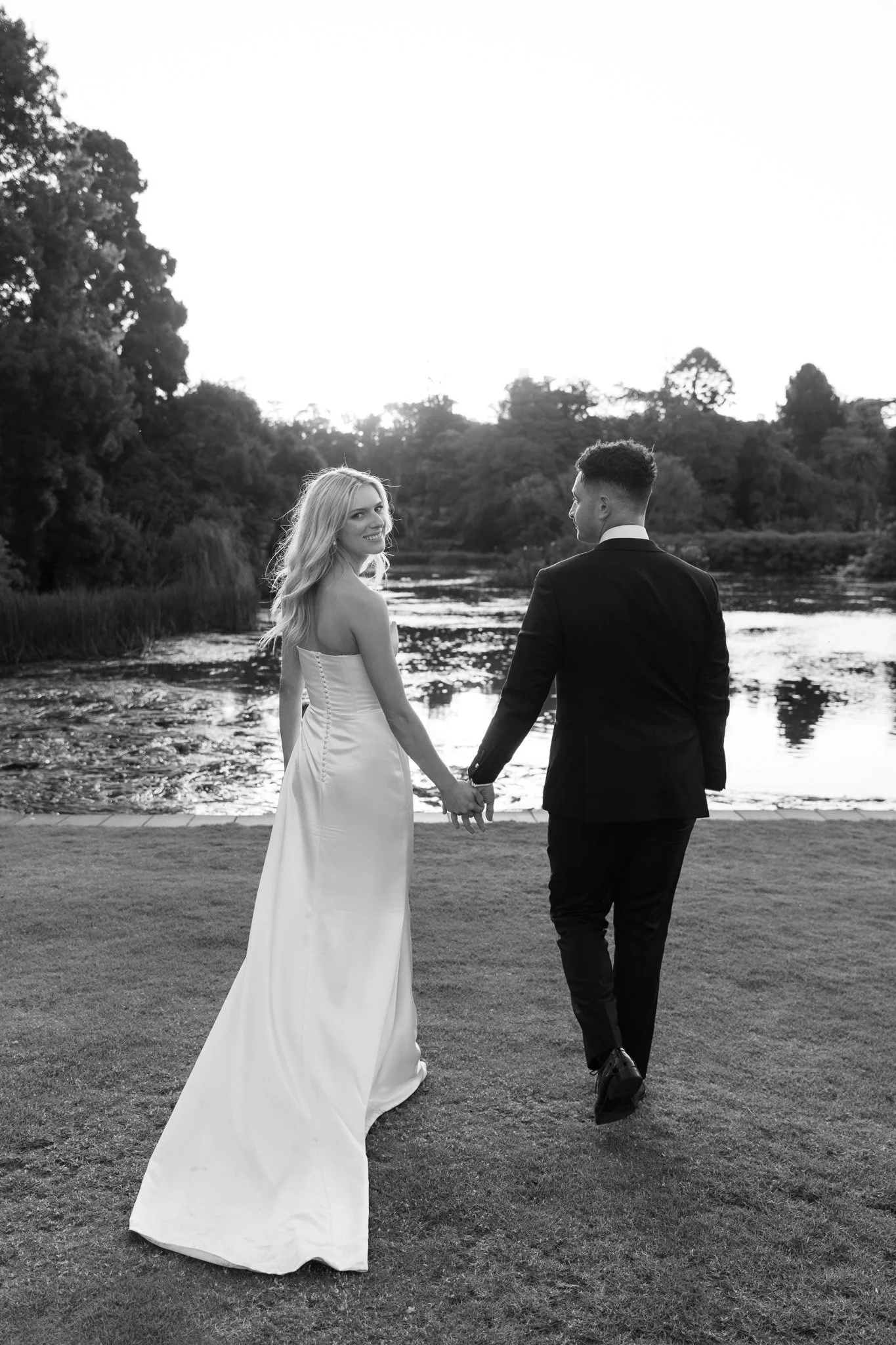 Black and white photo of a newlywed couple holding hands by a river, with trees in the background.