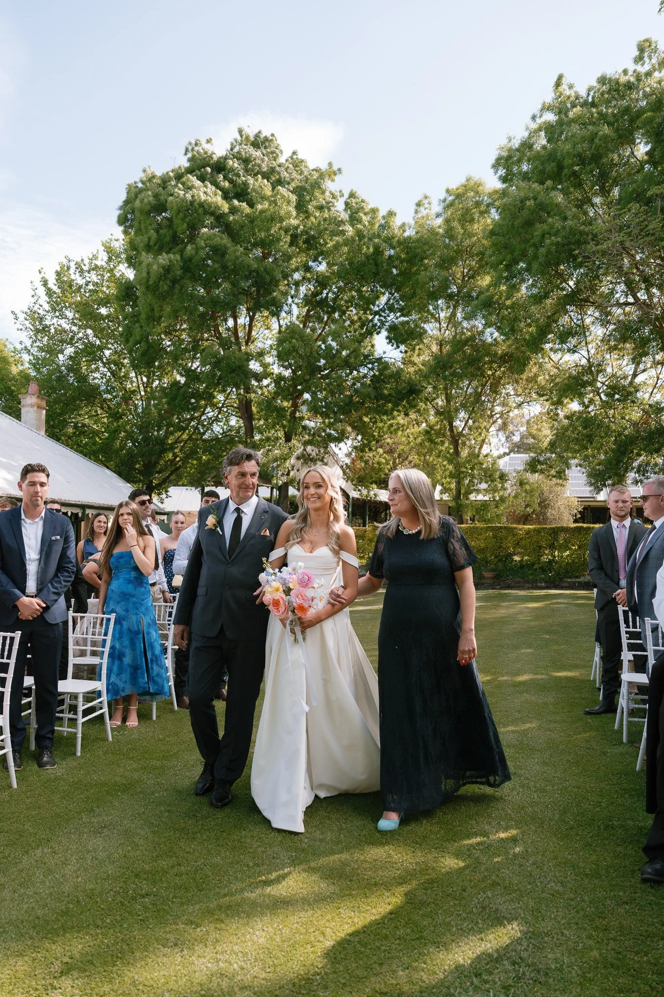 A bride walking down the aisle assisted by her mother, surrounded by seated wedding guests outdoors on a grassy lawn, with trees and a partly cloudy sky in the background.