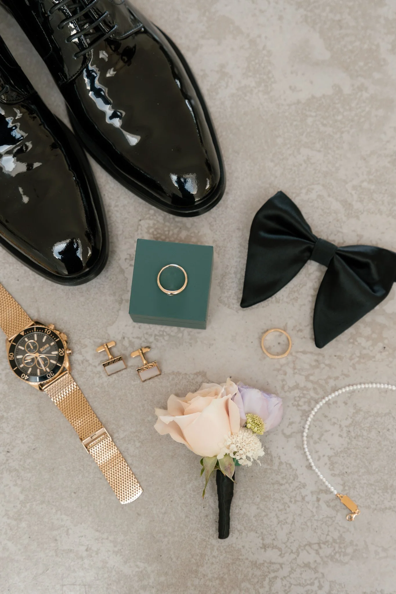 Black patent leather shoes, a green jewelry box with a silver ring, a black bow hair accessory, gold earrings, a gold wristwatch, a pearl necklace, a small bouquet of pink and white flowers, and a gold bracelet on a gray surface.