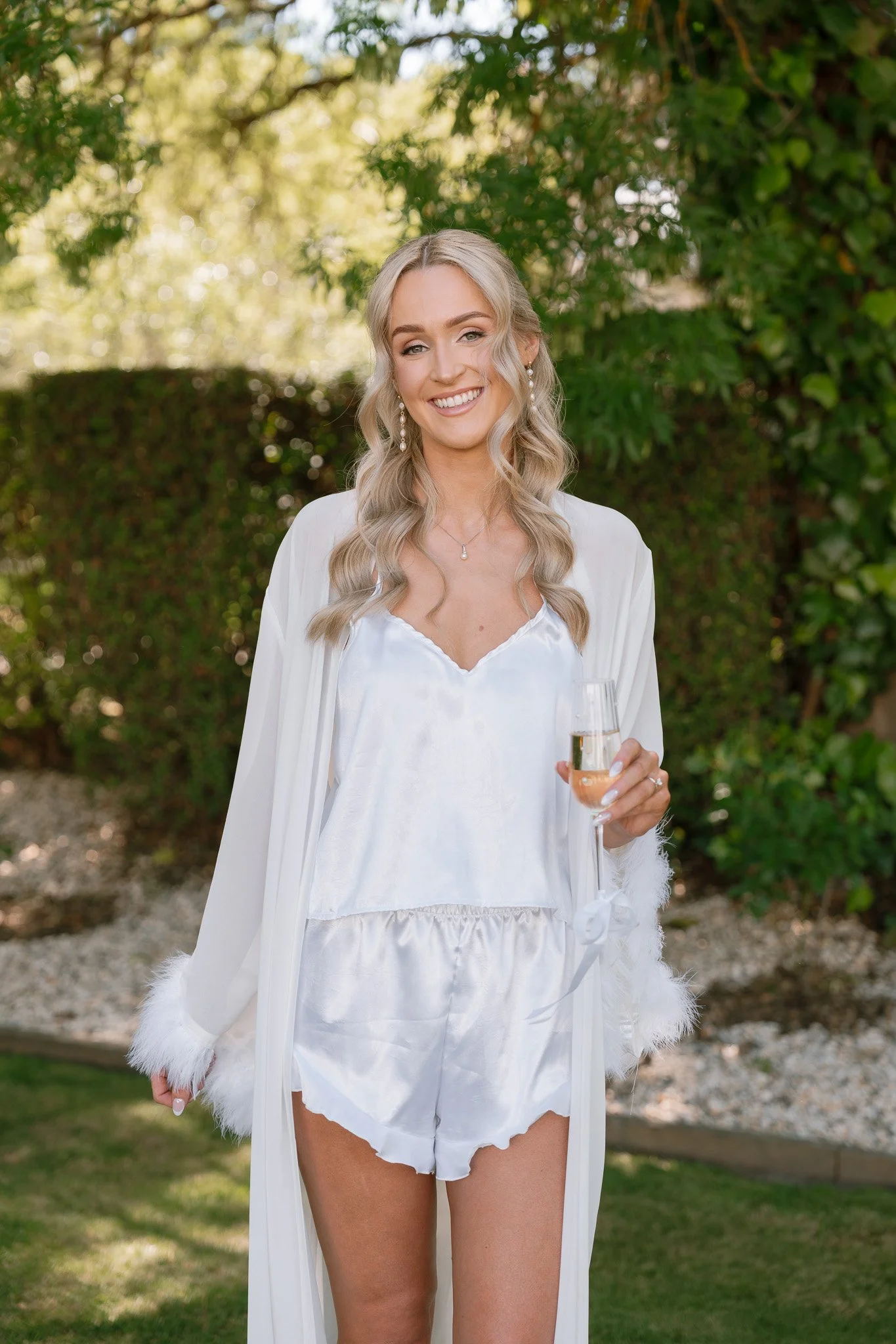 A woman in white satin pajamas and a long white robe with feathered cuffs, holding a glass of champagne, outdoors in a garden with trees and bushes.