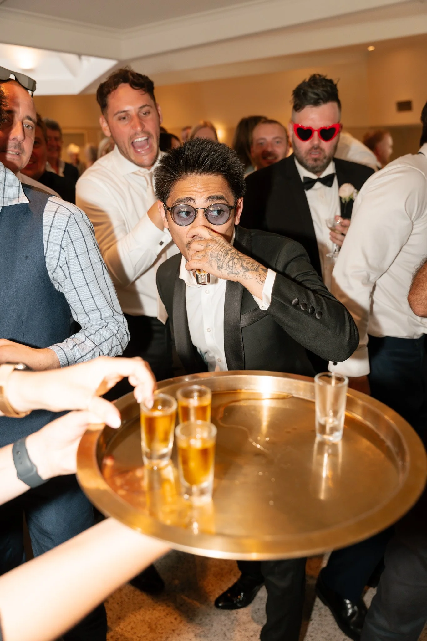 A group of people at a celebration or party, with some wearing sunglasses and dressed in formal attire. A person in the center, with tattoos and wearing dark sunglasses, is drinking from a shot glass while others around them are holding drinks and en