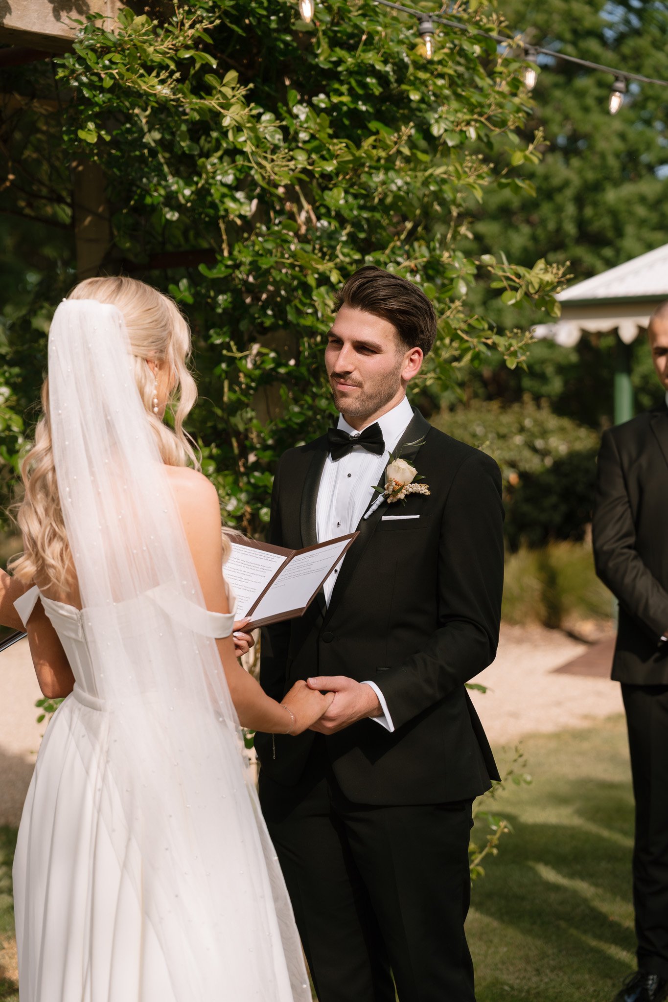 A bride and groom exchanging vows during an outdoor wedding ceremony, with a officiant reading from a book, surrounded by greenery.