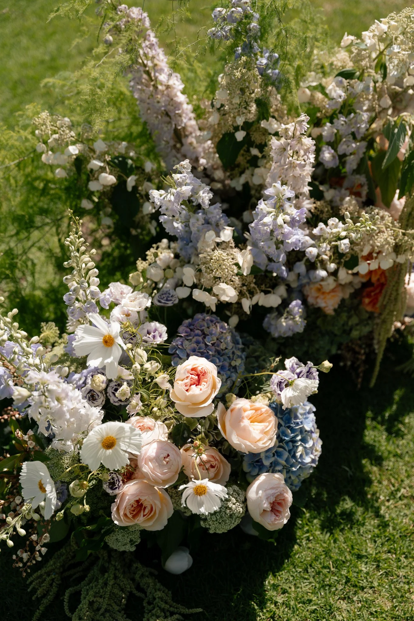A lush bouquet of flowers including light pink roses, white daisies, and blue hydrangeas, set against a grassy background.
