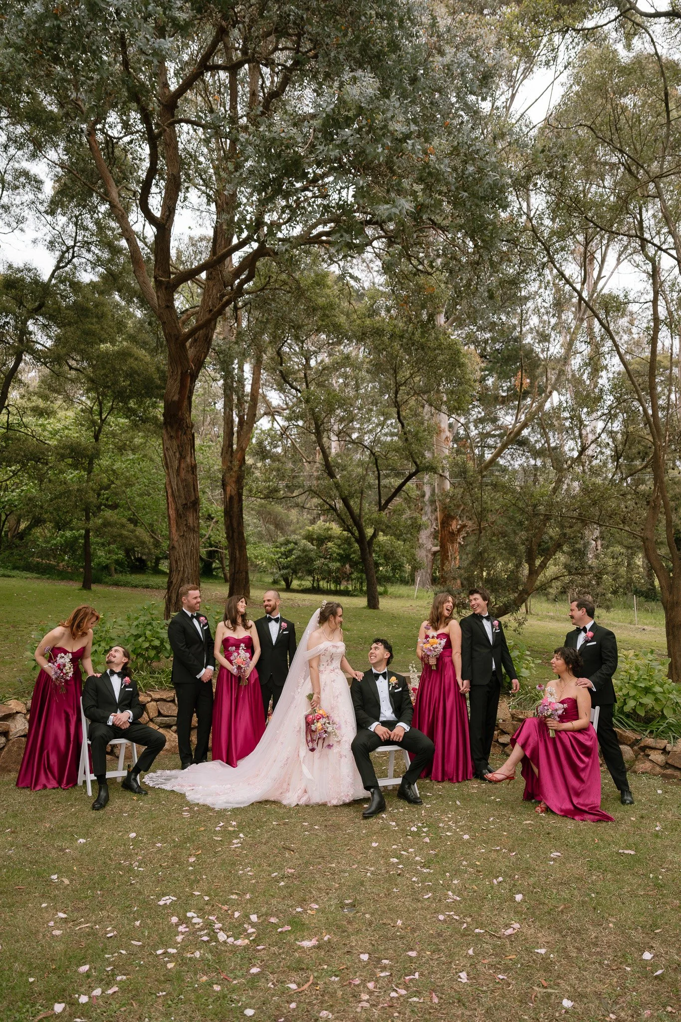 Wedding party outdoors among trees, bride in white gown with bouquet, groom in black suit, bridesmaids in matching magenta dresses, groomsmen in black suits, exchanging smiles and laughter.
