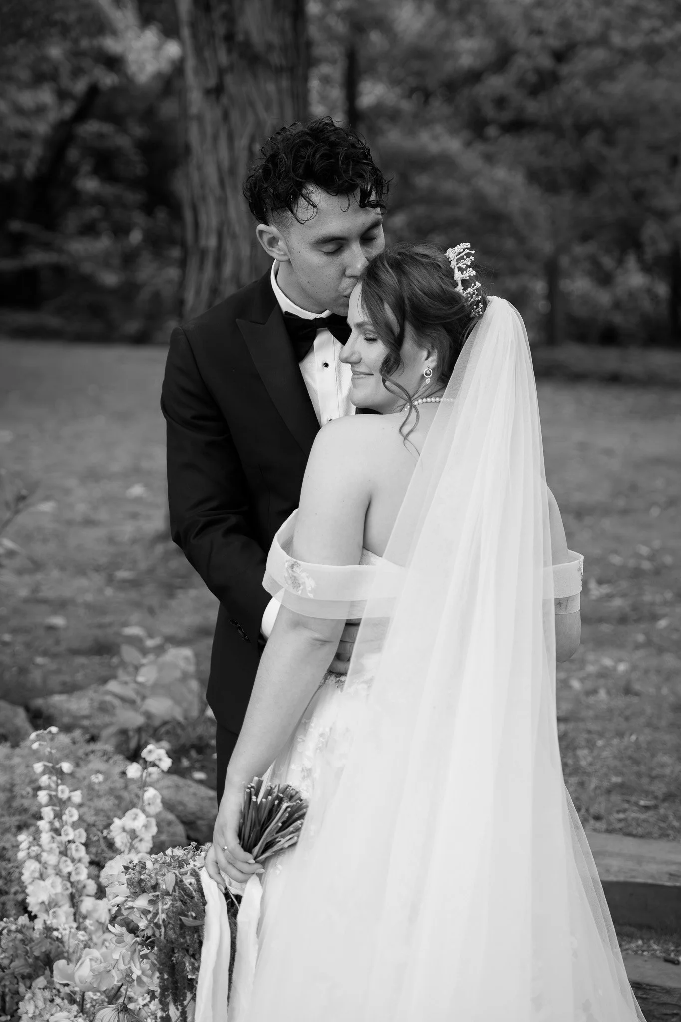 A black and white photo of a bride and groom on their wedding day. The groom, dressed in a tuxedo, is kissing the bride's forehead. The bride, in a wedding dress with a veil, is holding a bouquet of flowers and smiling contentedly.