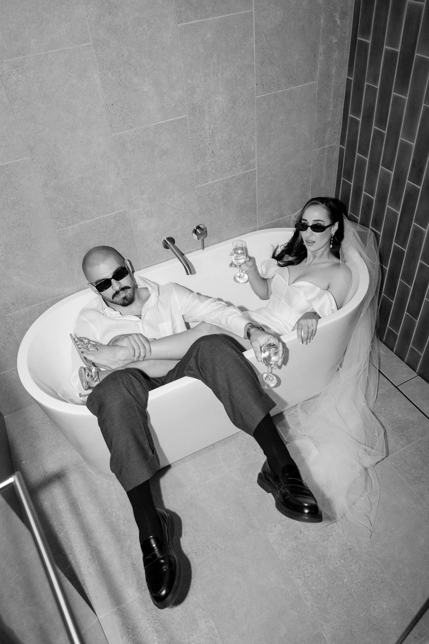 A man and woman wearing sunglasses sitting in a bathtub with champagne glasses. The woman is in a wedding dress with a veil, and the man is dressed in formal attire. The scene appears staged for a photo.