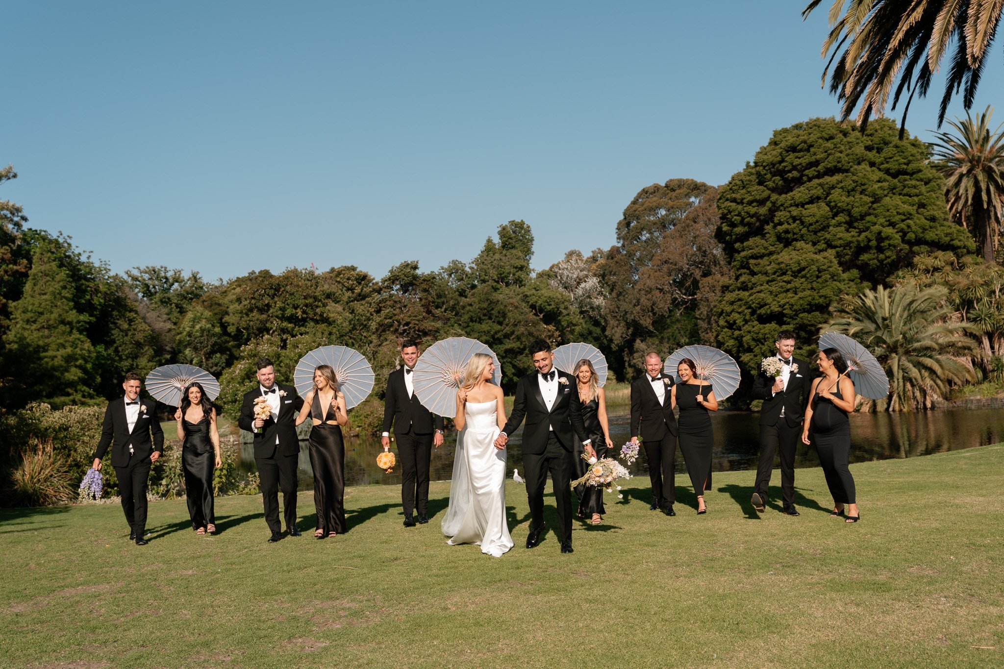 A wedding party walking across a grassy area by a pond, with trees and blue sky in the background. The bride and groom are in the center, holding hands, with bridesmaids and groomsmen on either side, some holding umbrellas.