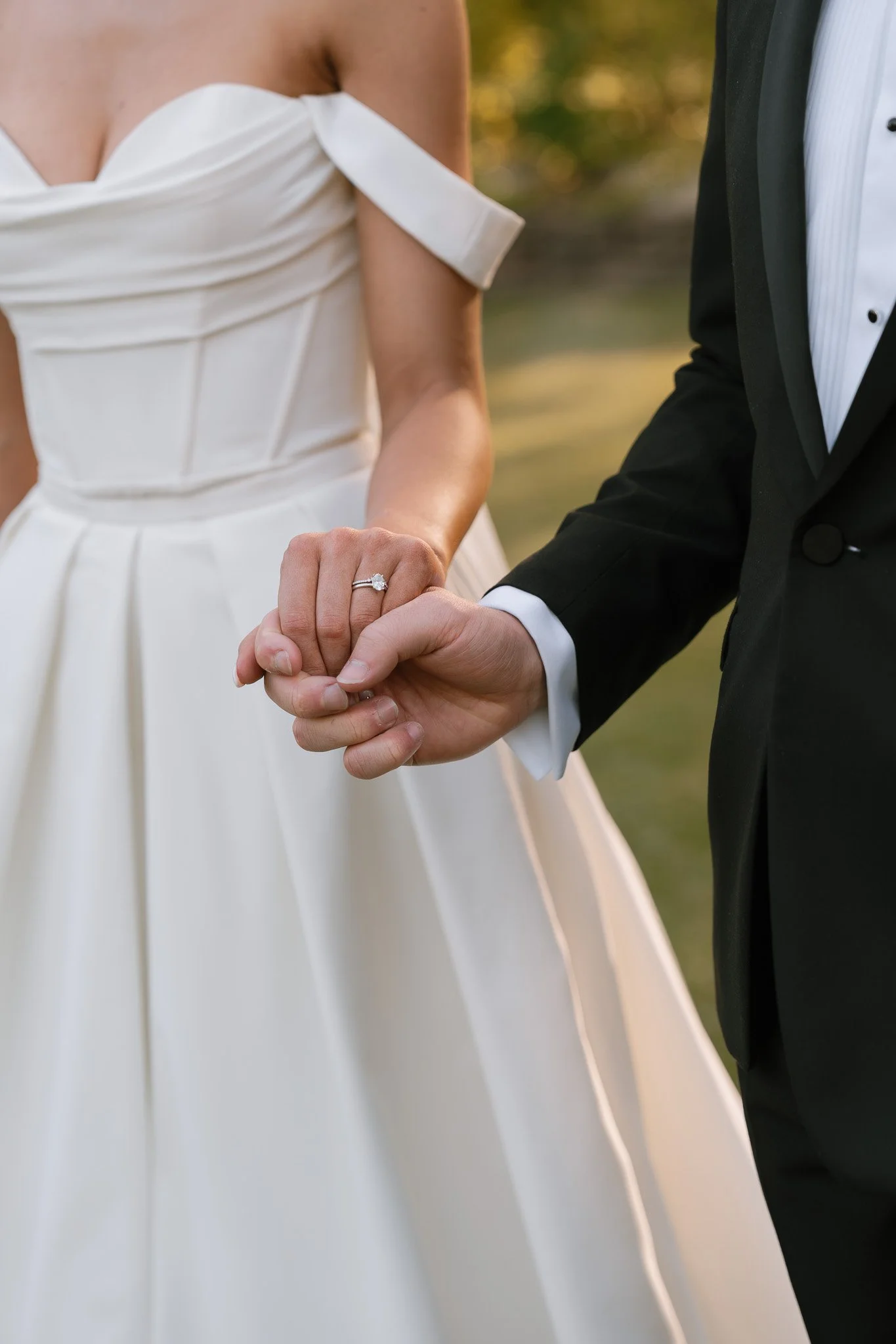Close-up of a bride and groom holding hands, showing the bride's wedding ring, during a wedding ceremony outdoors.