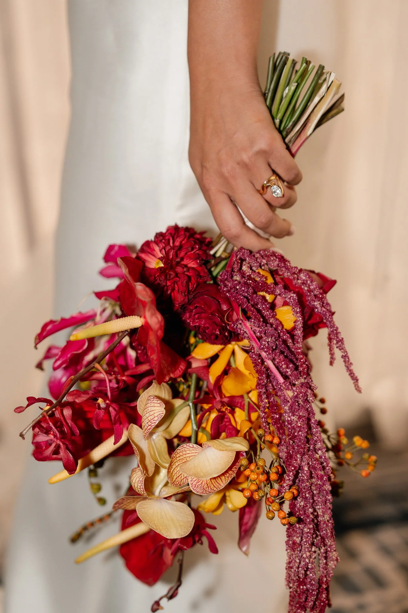 A person in a white dress holding a colorful bouquet of flowers with a gold ring on their finger.