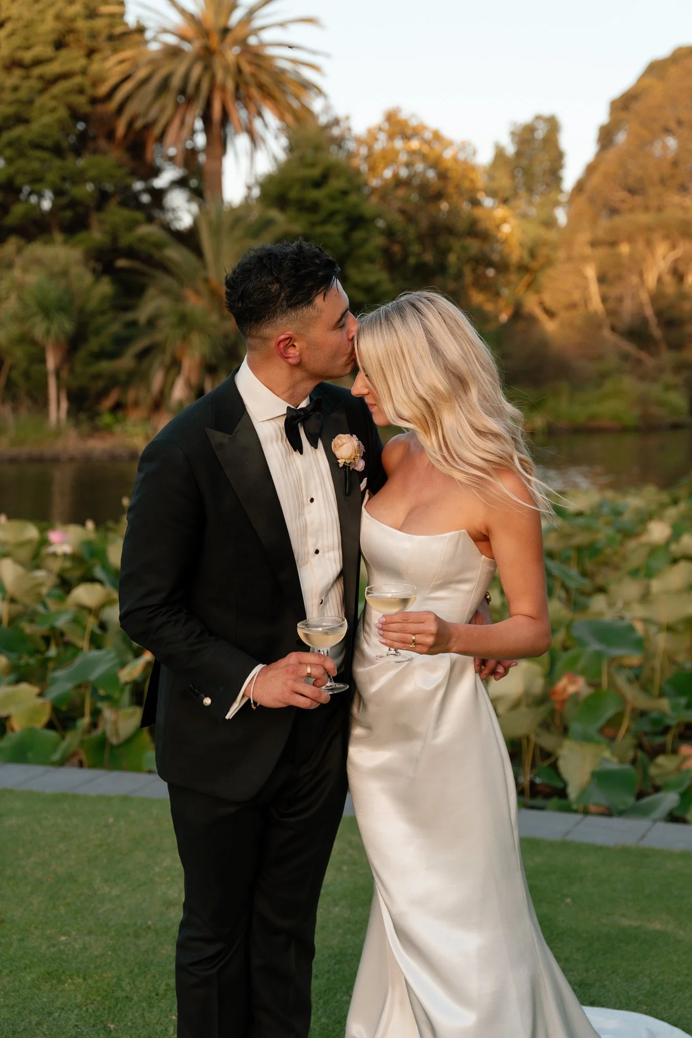 A newlywed couple with light skin and slender physiques, dressed in formal wedding attire, sharing a tender moment outdoors by a pond with lush green foliage and trees in the background, during sunset.