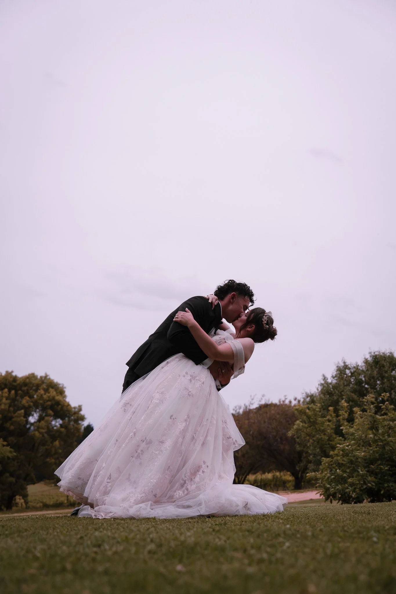A couple in wedding attire sharing a kiss outdoors, with the groom holding the bride and the bride in a white wedding dress, tall grass, and trees in the background under an overcast sky.