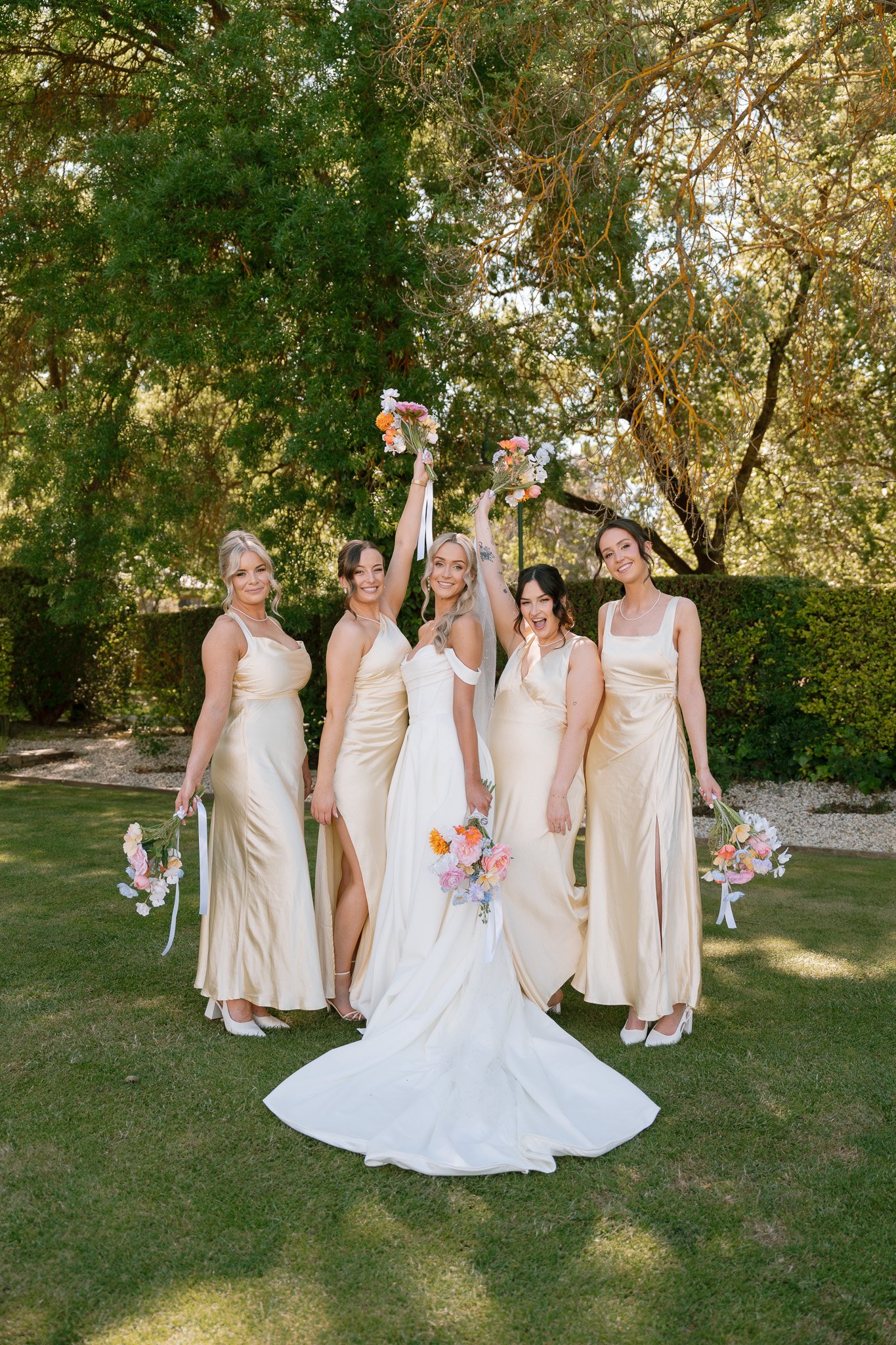A group of women, including a bride in a white wedding dress and five bridesmaids in matching gold dresses, celebrating outdoors with bouquets of flowers under a large tree.