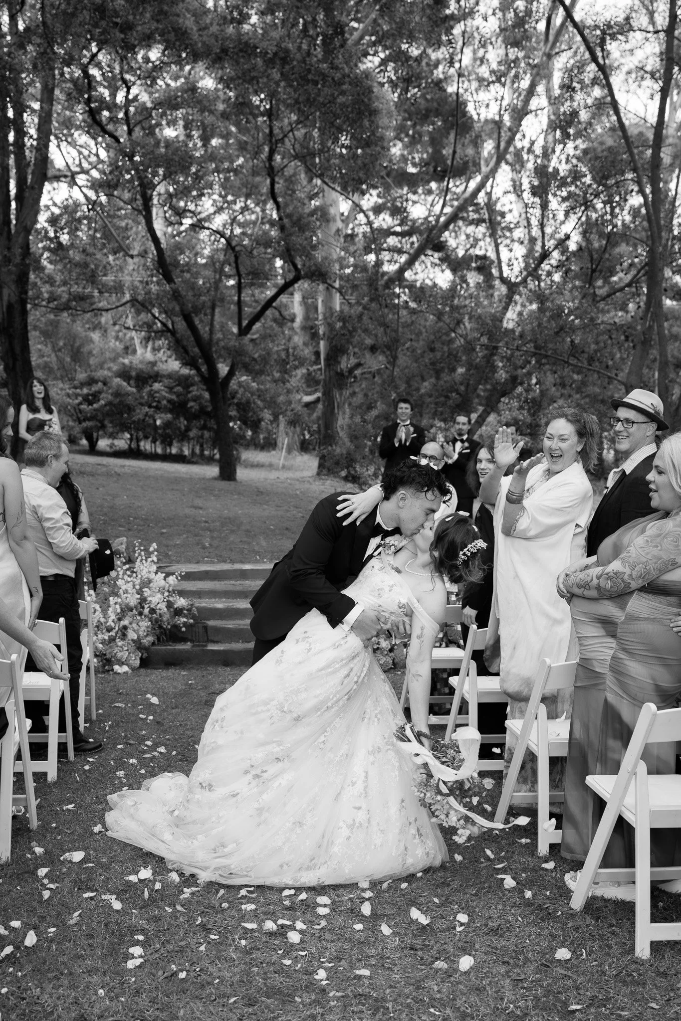 Black and white photo of a wedding scene outdoors, showing a bride and groom sharing a kiss and the bride bending forward. Guests are clapping and smiling, some standing and some sitting, with trees in the background.