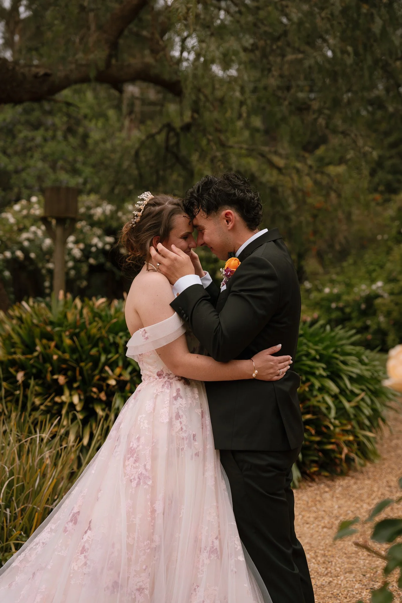 A newlywed couple sharing a romantic moment outdoors, with their foreheads touching and eyes closed, surrounded by lush greenery.