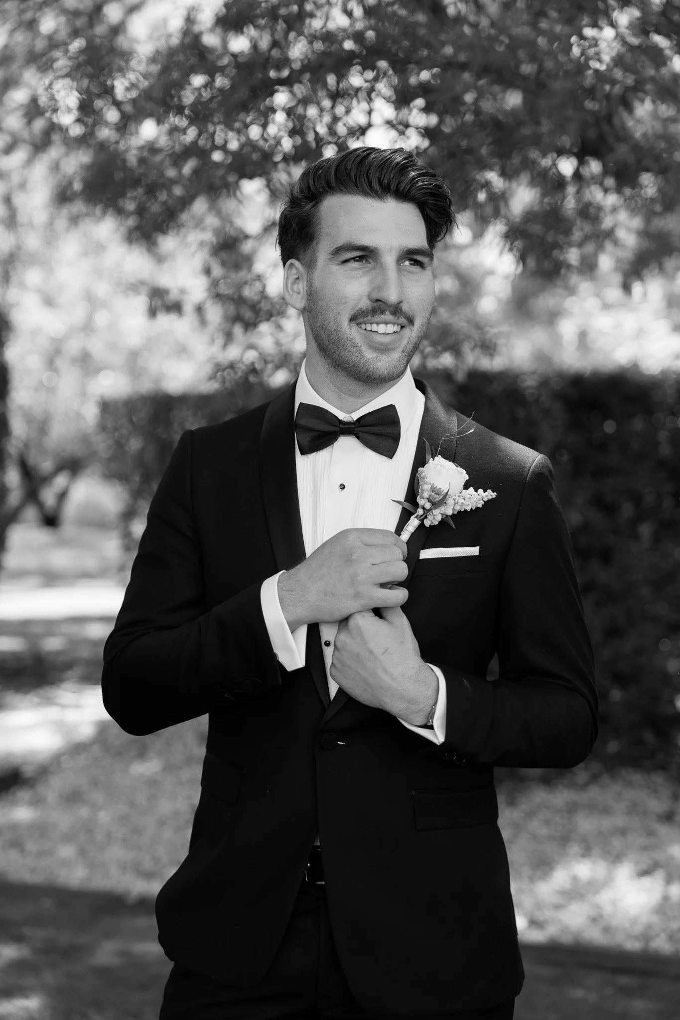 A man dressed in a tuxedo with a bow tie, holding a boutonnière, smiling outdoors with trees in the background.