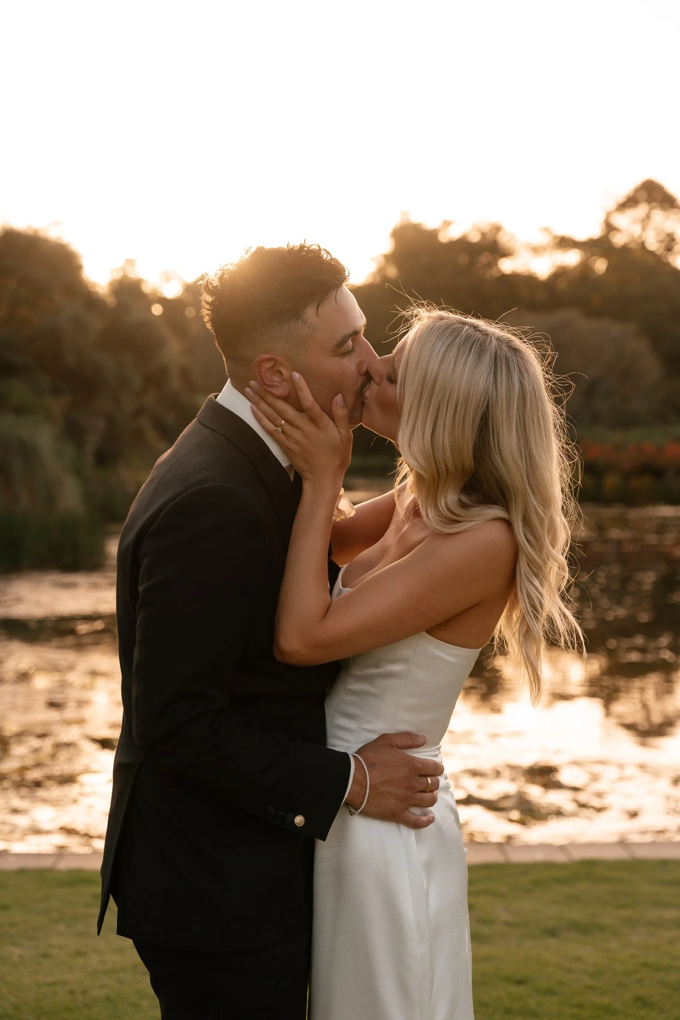 A couple in wedding attire kissing outdoors during sunset, with water and trees in the background.