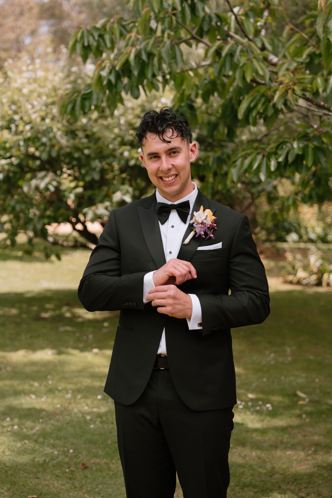 A young man in a black tuxedo with a white shirt and a black bow tie, standing outdoors in a park, smiling and holding his hands together. He has a boutonniere on his left lapel, with pink and purple flowers, and is surrounded by greenery.