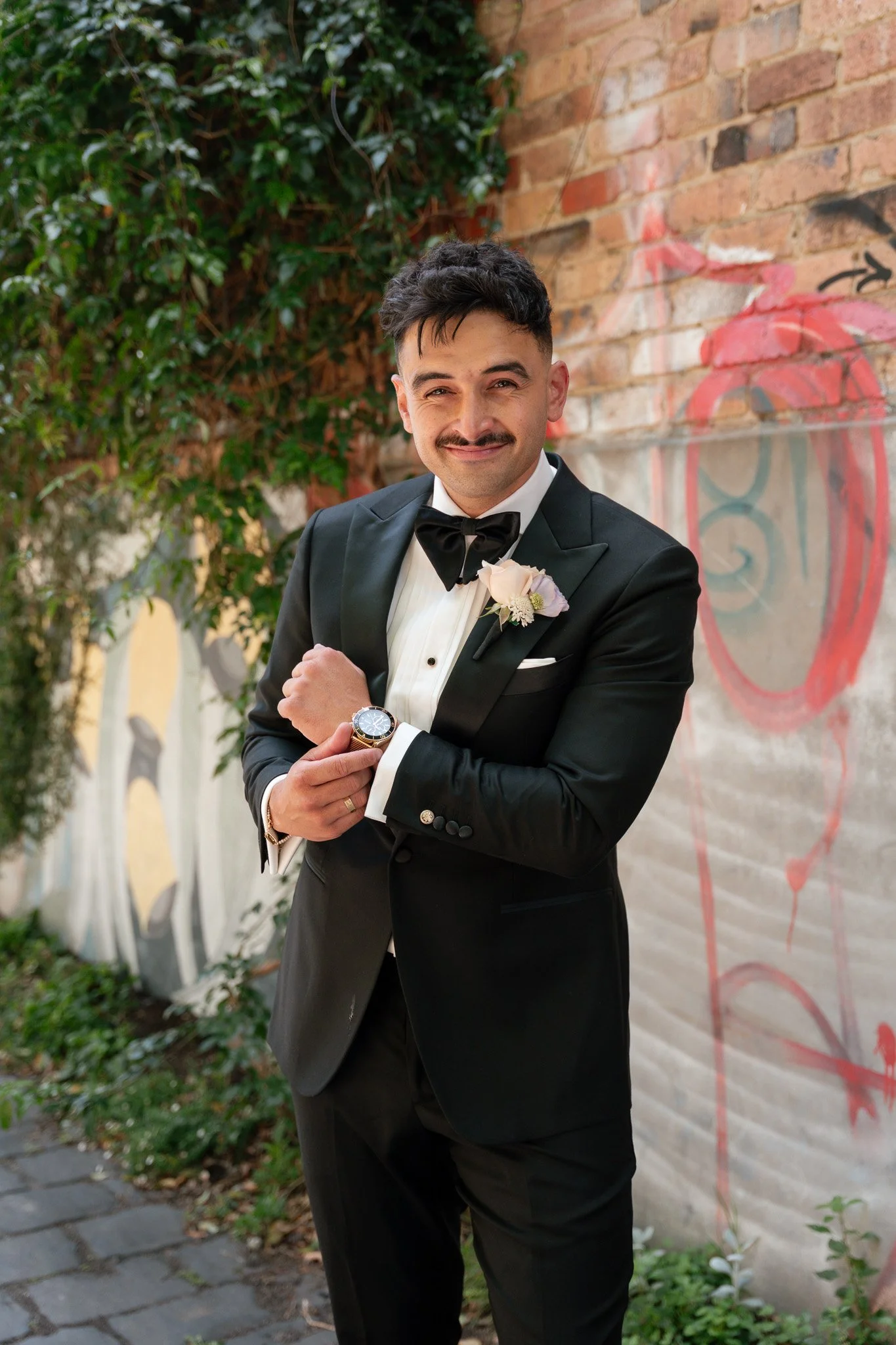 A man in a tuxedo with a boutonniere, smiling and looking confident, standing outdoors near a brick wall with graffiti and greenery.