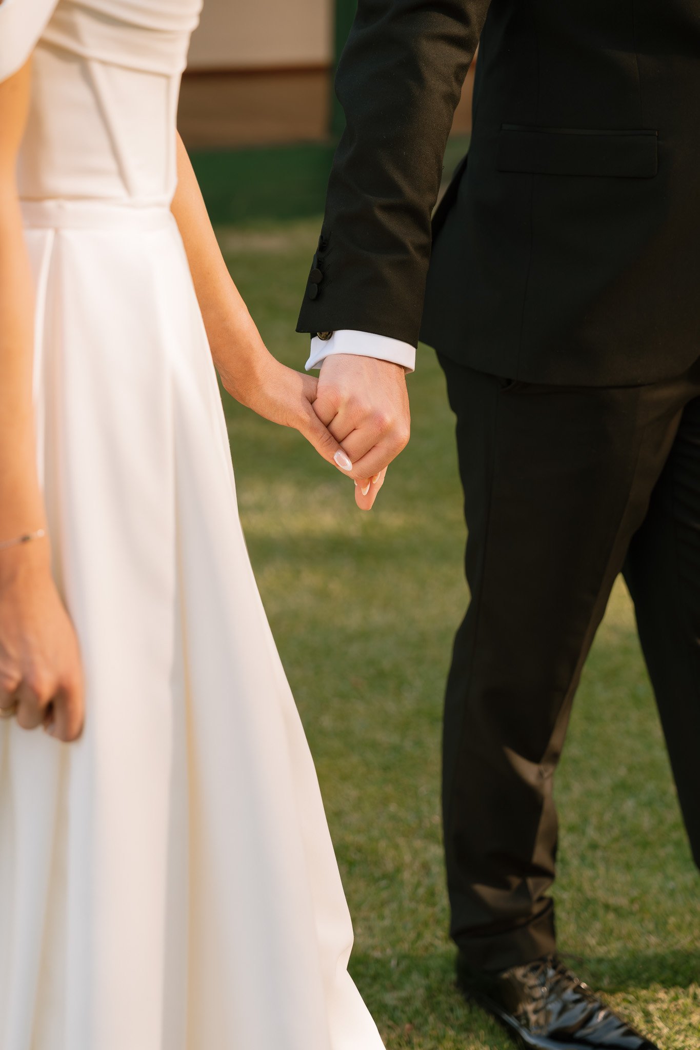 Close-up of a couple holding hands, dressed in wedding attire, with the woman wearing a white wedding dress and the man in a black suit, outdoors on grass.