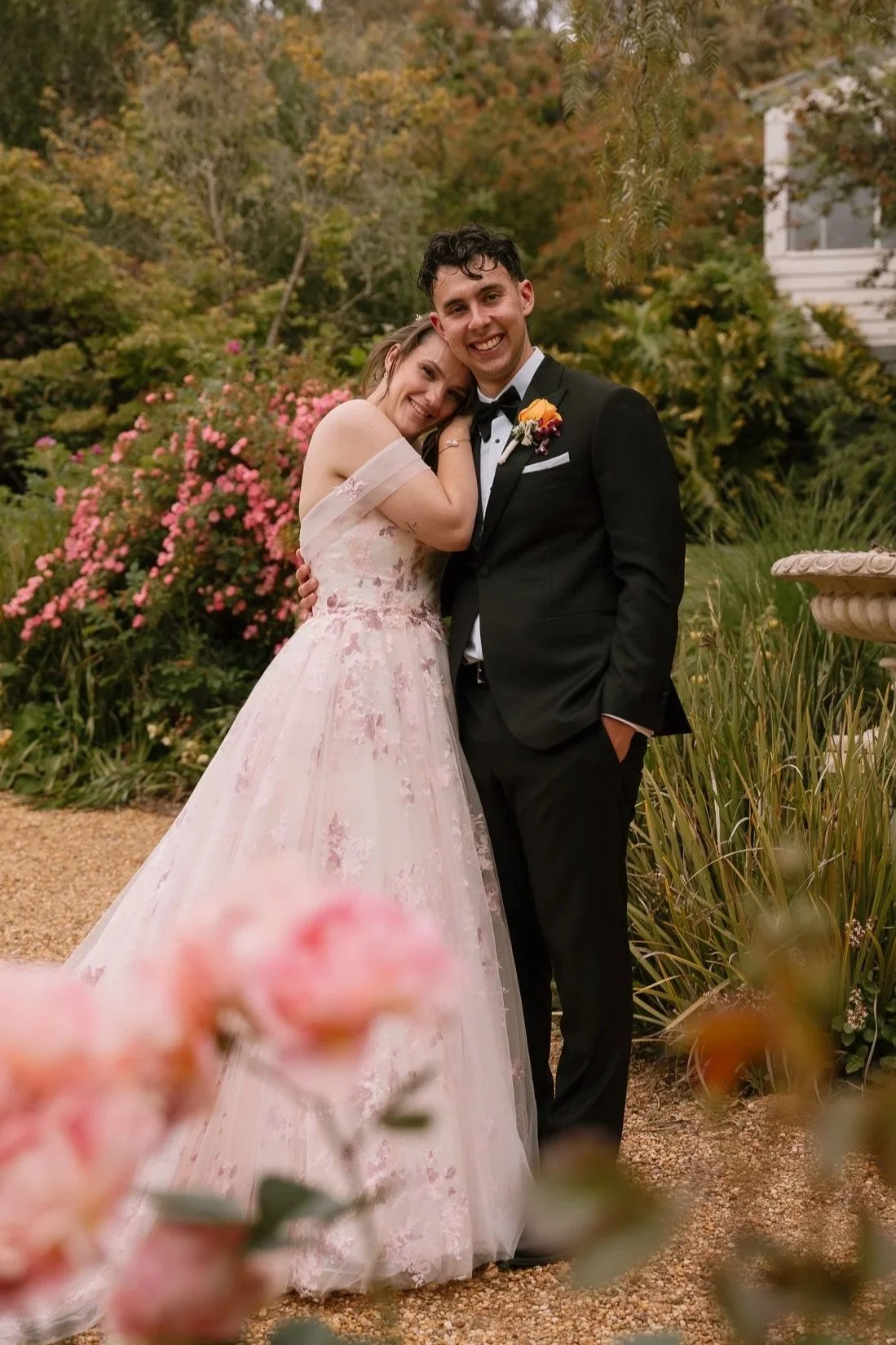 A bride and groom smiling and embracing in a garden during their wedding, with colorful flowers and foliage in the background.