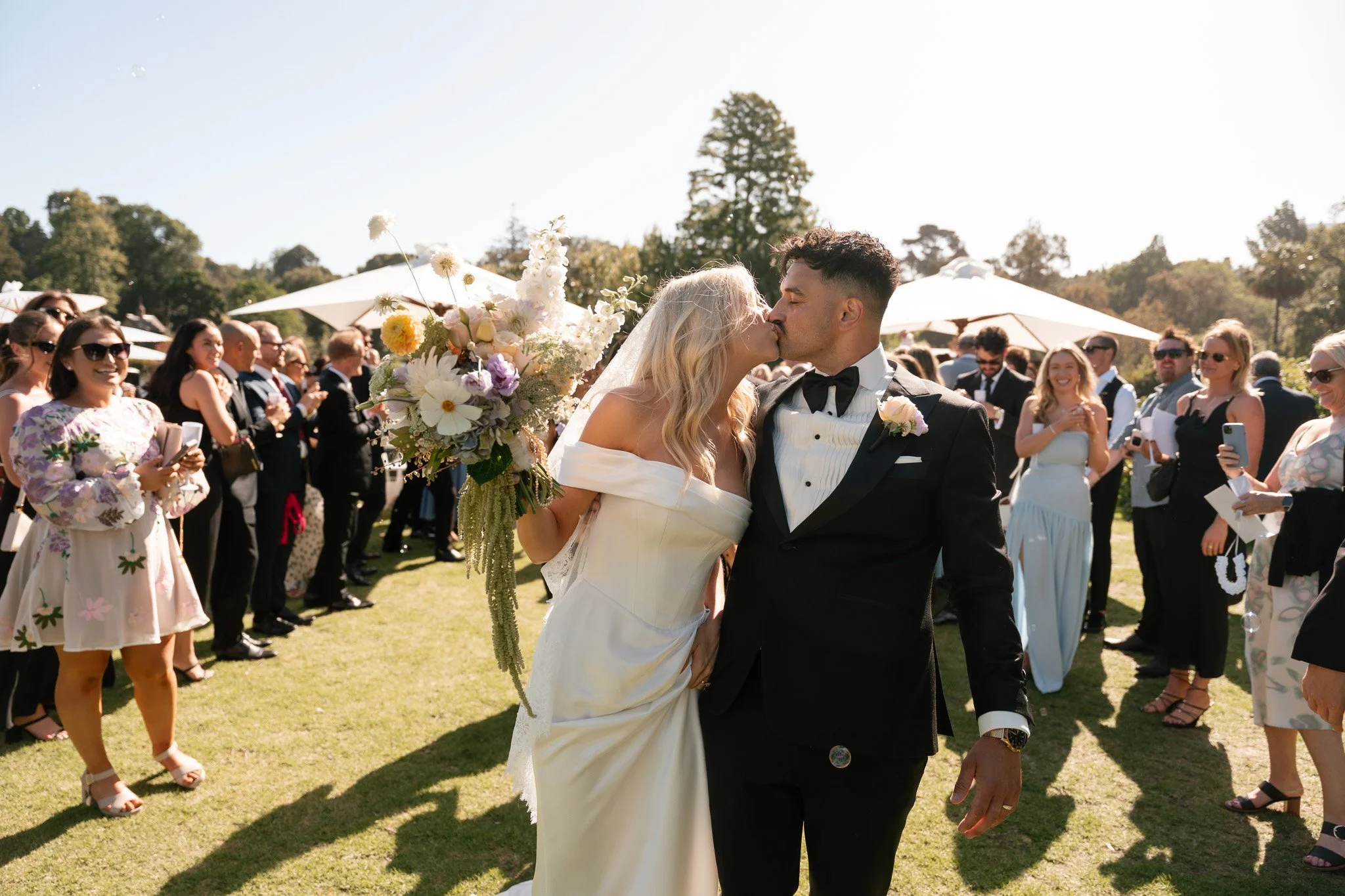 A newlywed couple sharing a kiss outdoors at their wedding reception surrounded by guests, with the bride holding a large floral bouquet and wearing an off-shoulder white wedding gown, and the groom in a black tuxedo with a bowtie, on a sunny day wit