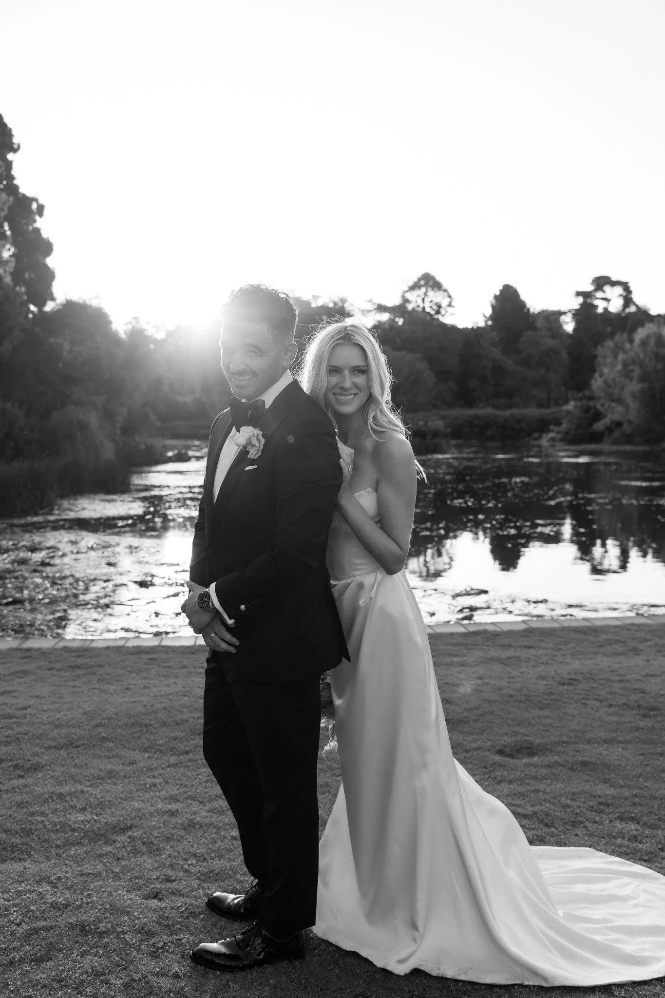 A black and white photo of a smiling bride and groom standing outdoors near a river at sunset.