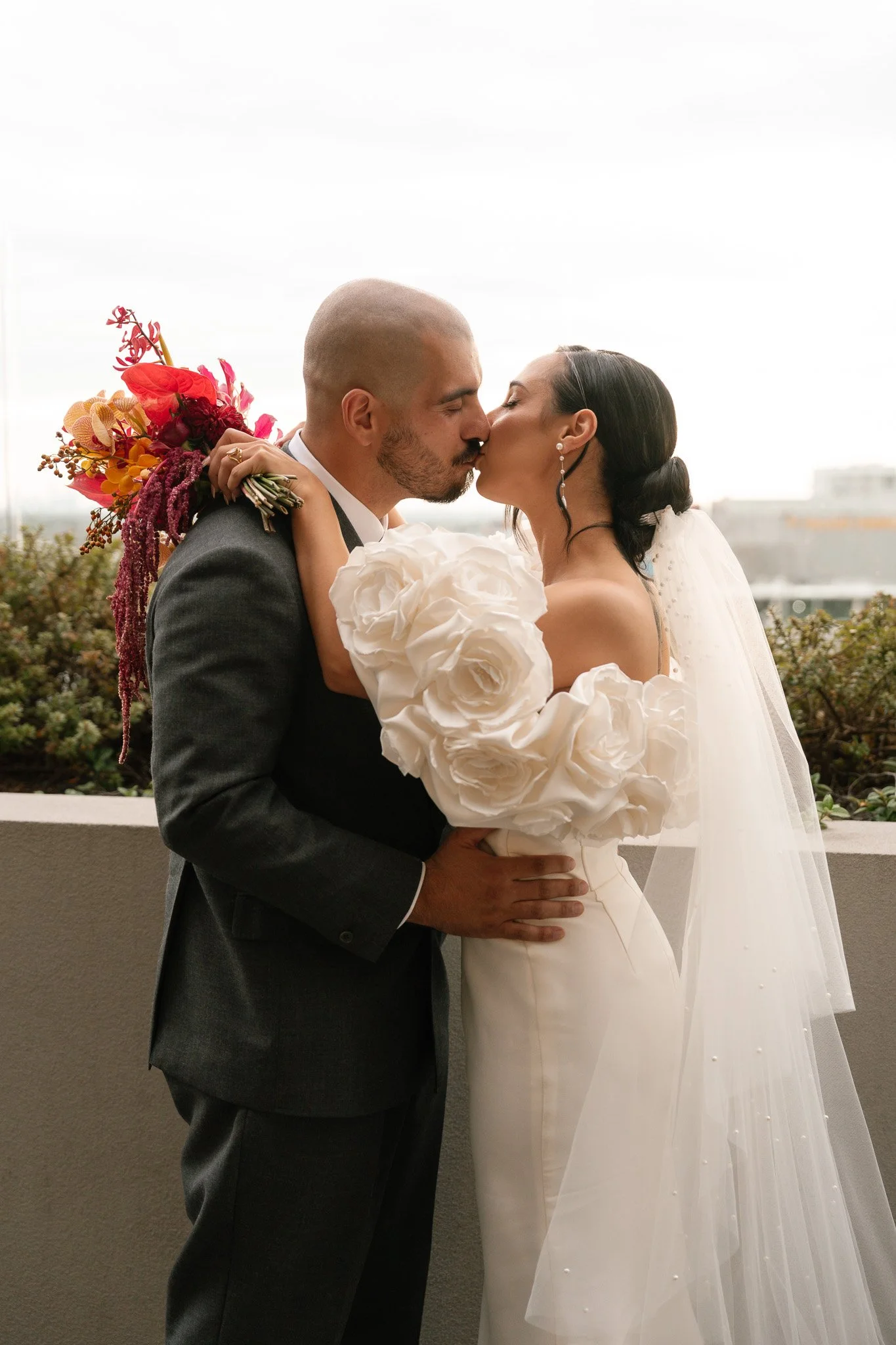 Couple in wedding attire, embracing and kissing on a balcony, with cityscape background and bouquet.