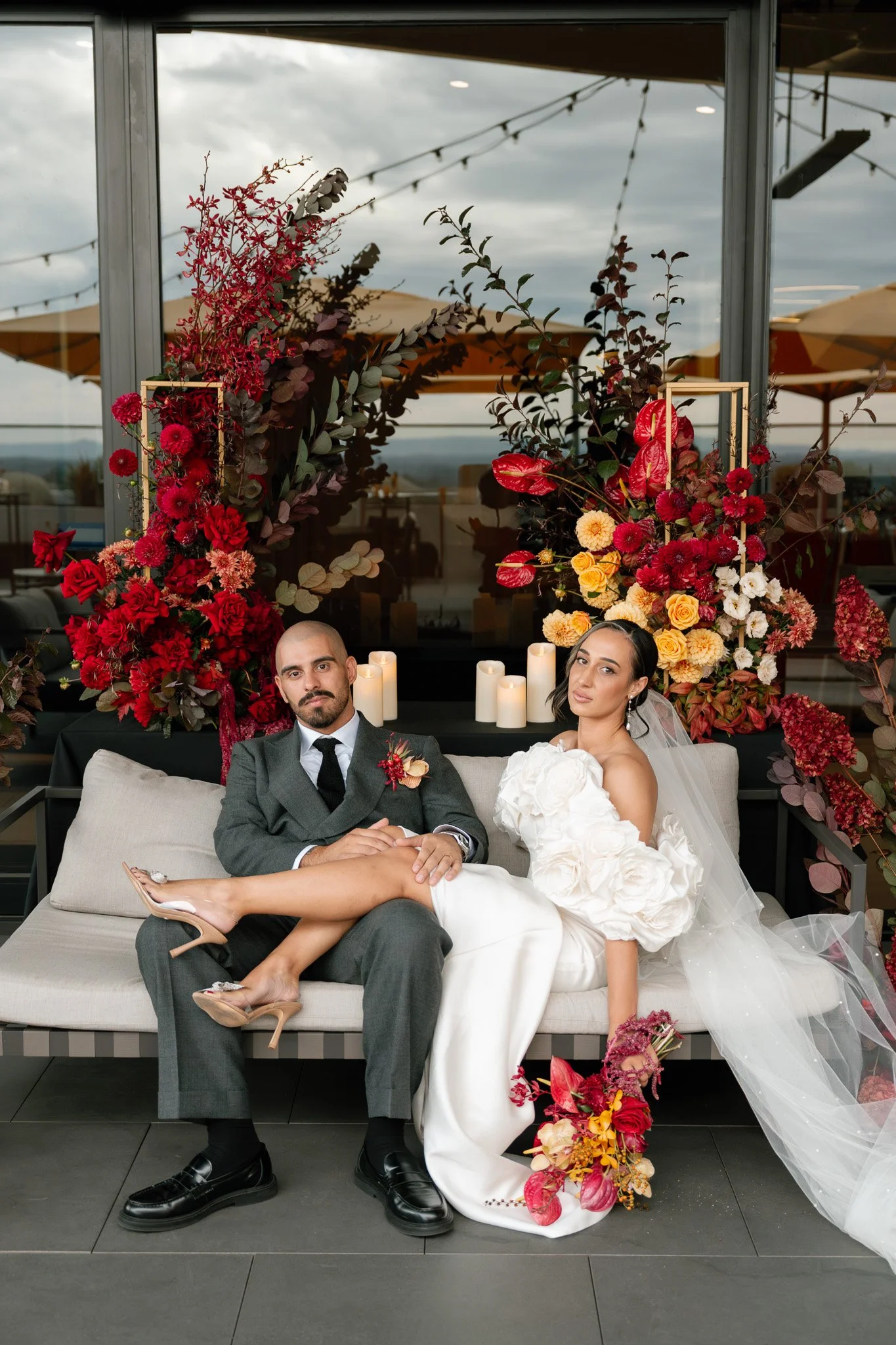 A wedding couple sitting on a sofa with floral and candle decorations behind them. The groom is wearing a gray suit, and the bride is in a white wedding dress with floral details, holding a bouquet of flowers.