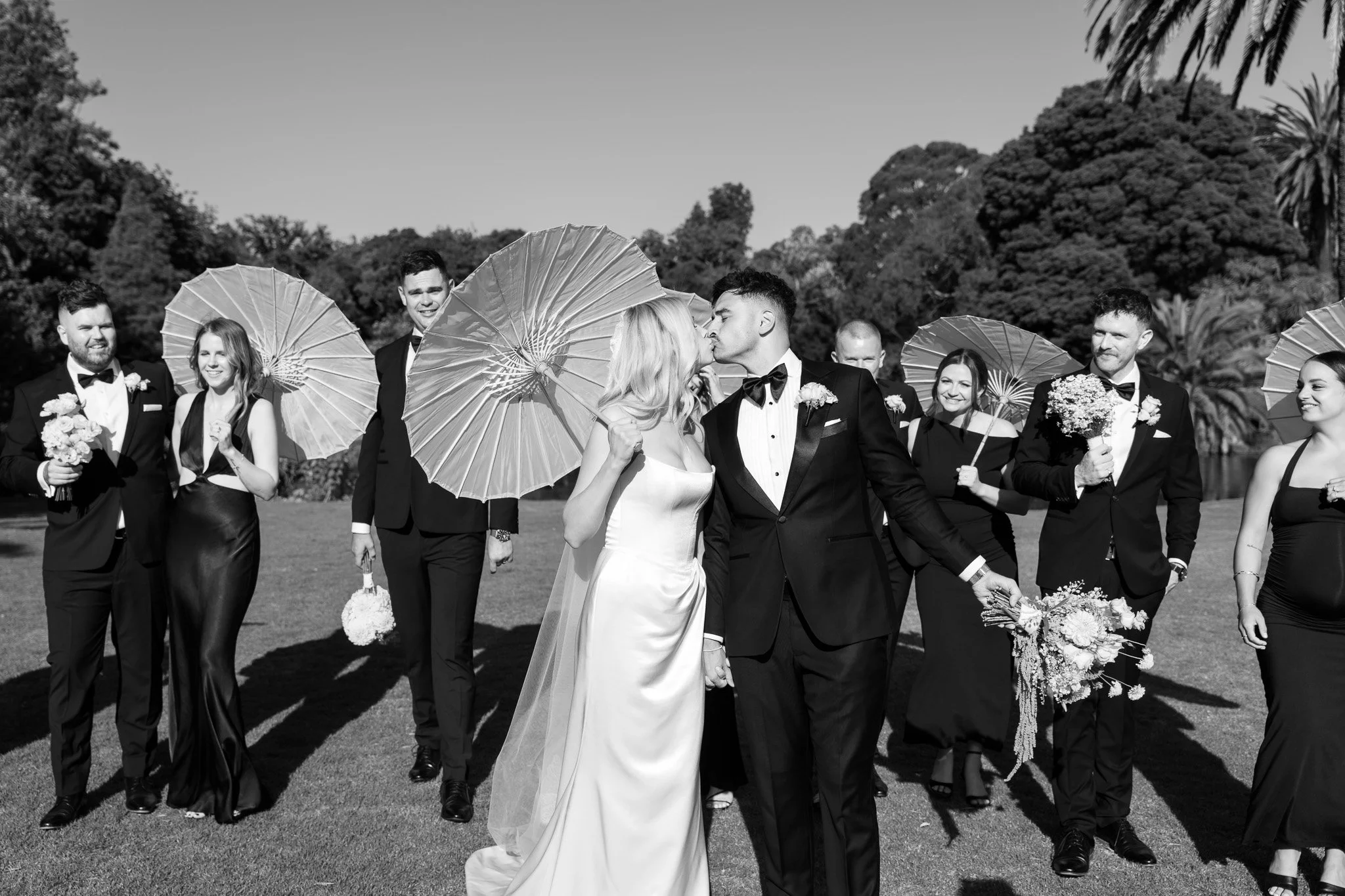 Black and white wedding photo of a couple kissing, surrounded by bridal party holding umbrellas and flowers outdoors.