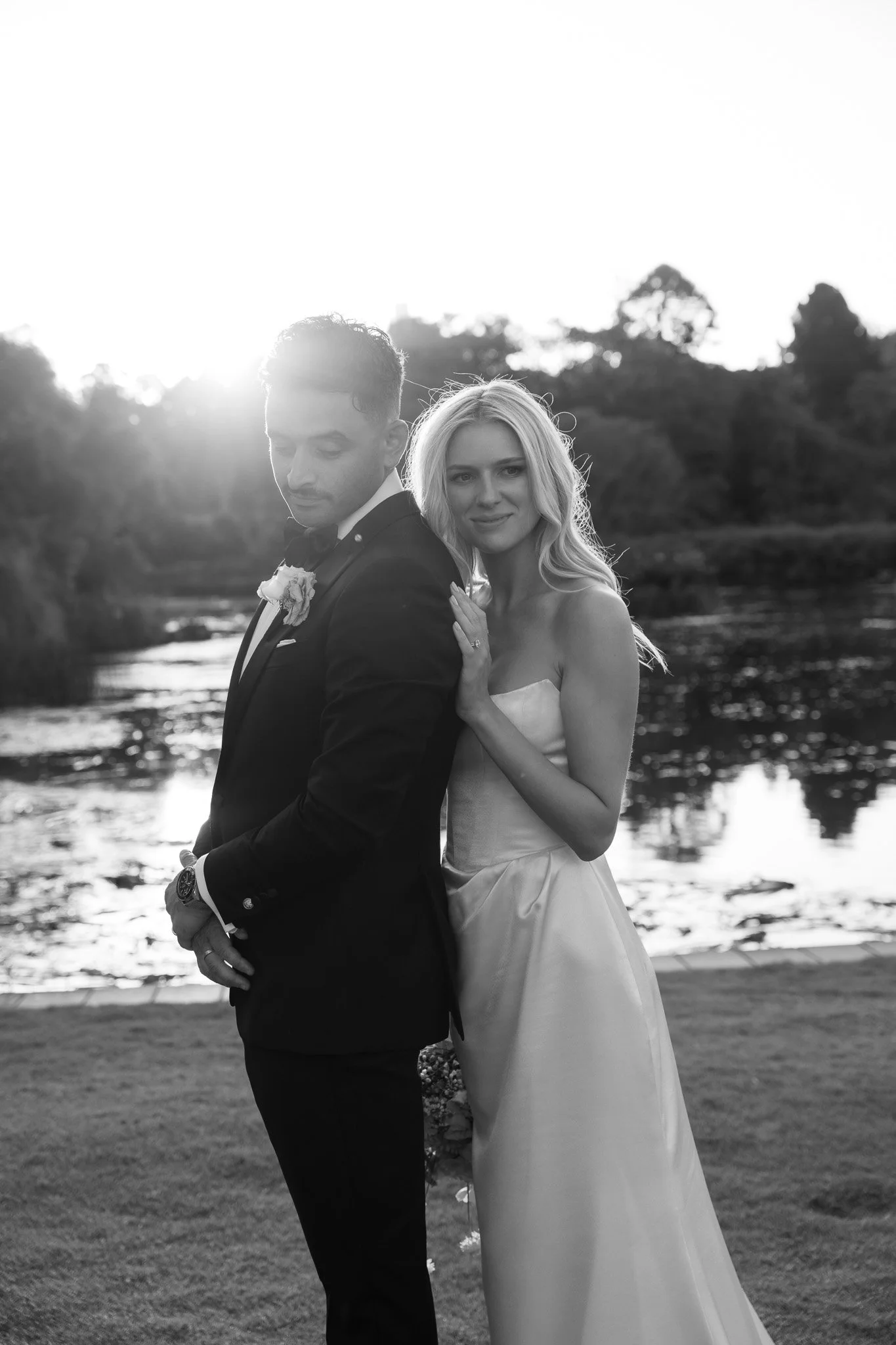 Black and white photo of a newlywed couple standing outdoors near a river at sunset. The groom is dressed in a tuxedo with a boutonniere, and the bride is in a strapless wedding gown with loose wavy hair. The bride gently touches the groom's shoulder