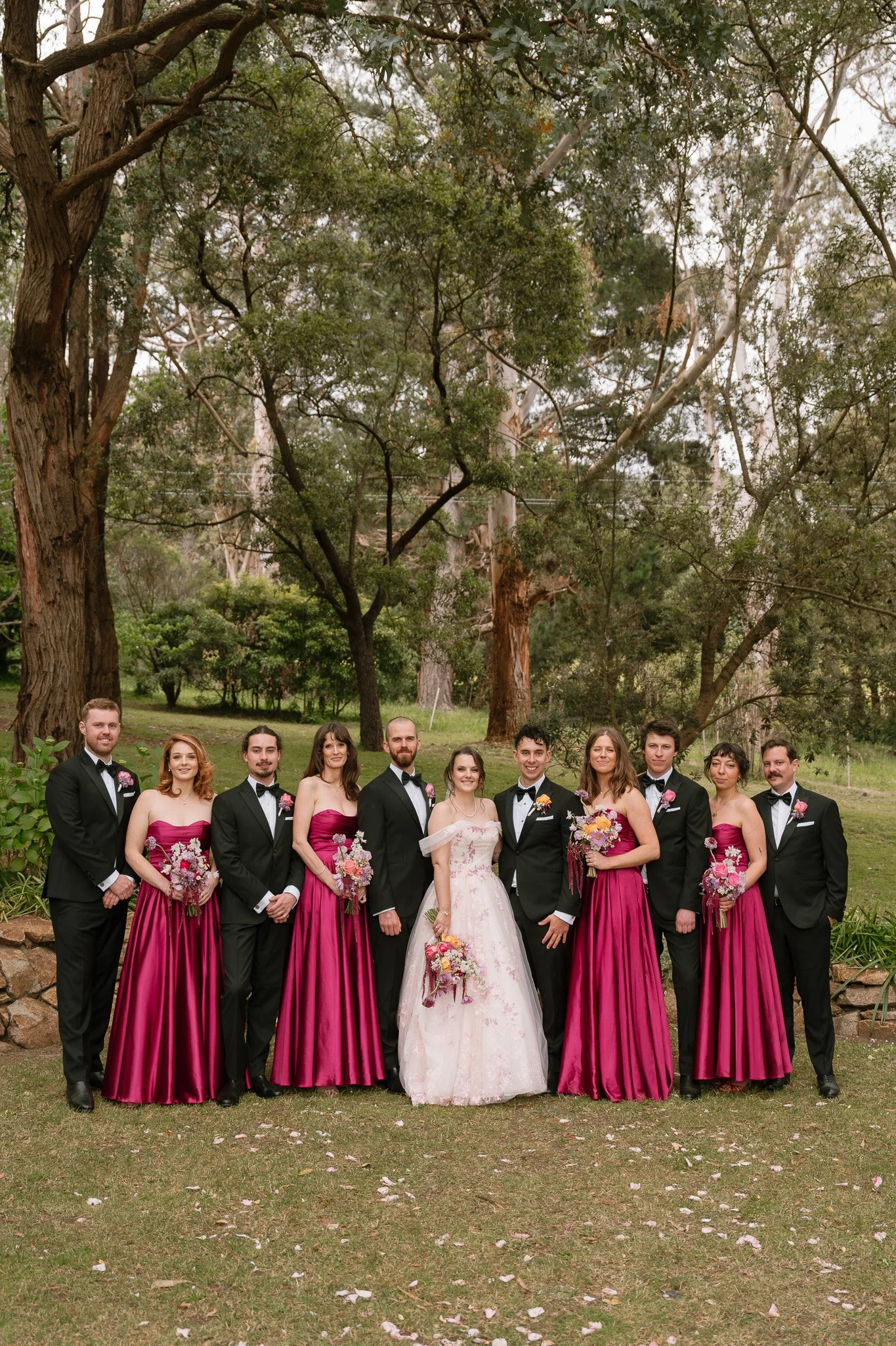 Wedding party outdoors, group of ten people, including bride in white dress holding a bouquet, groom in black tuxedo, and bridesmaids in pink dresses holding bouquets, standing on grass with trees in background.