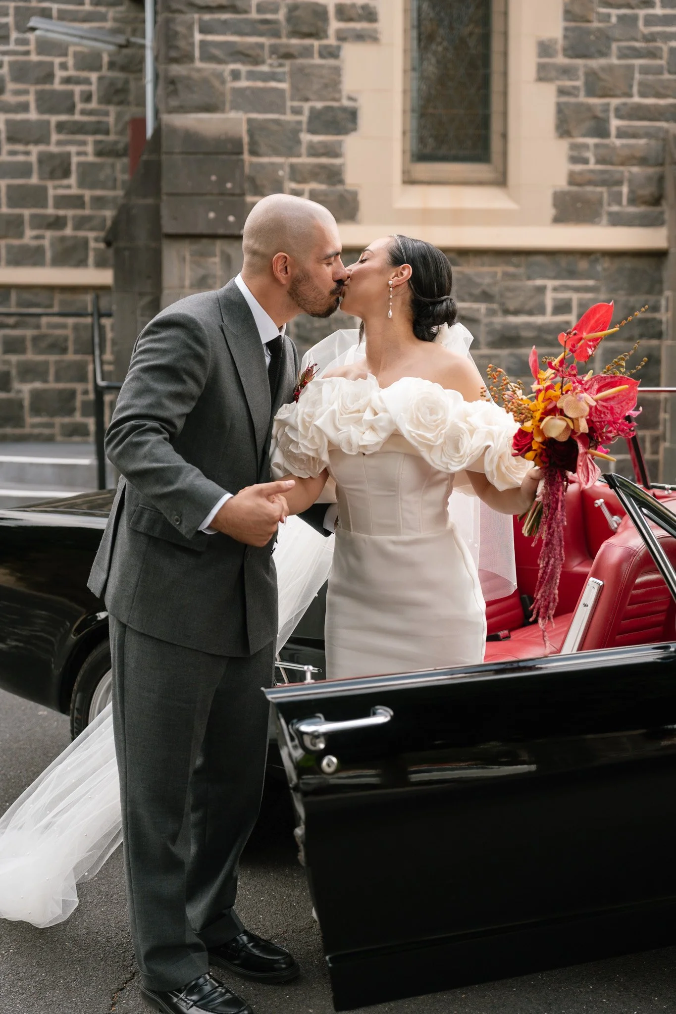 A newlywed couple sharing a kiss beside a vintage car decorated with a bouquet of flowers, with a stone church in the background.