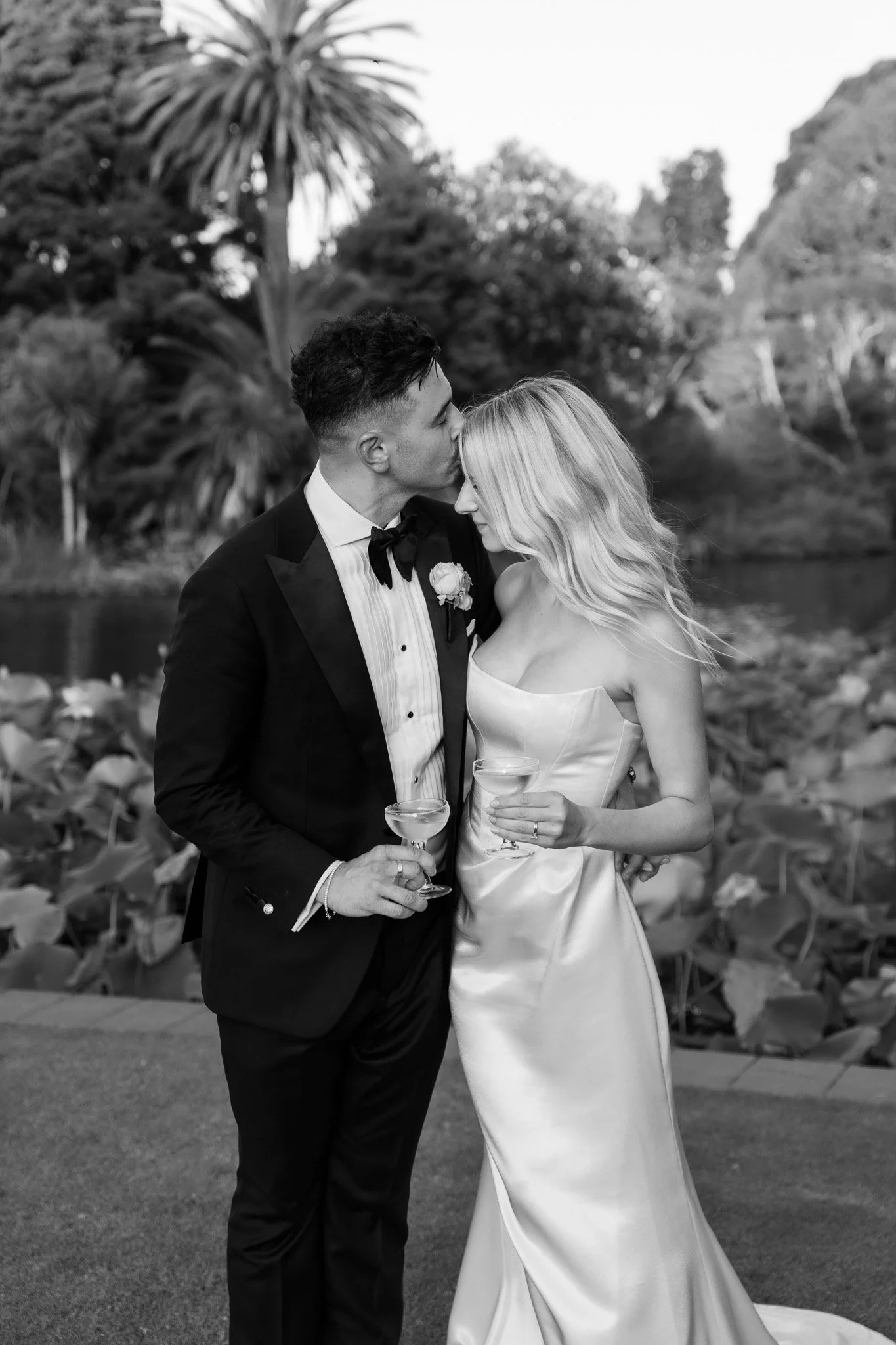 Black and white photo of a newlywed couple, the groom kissing the bride on her forehead, both holding champagne glasses, outdoors near a lake with trees and rocks in the background.