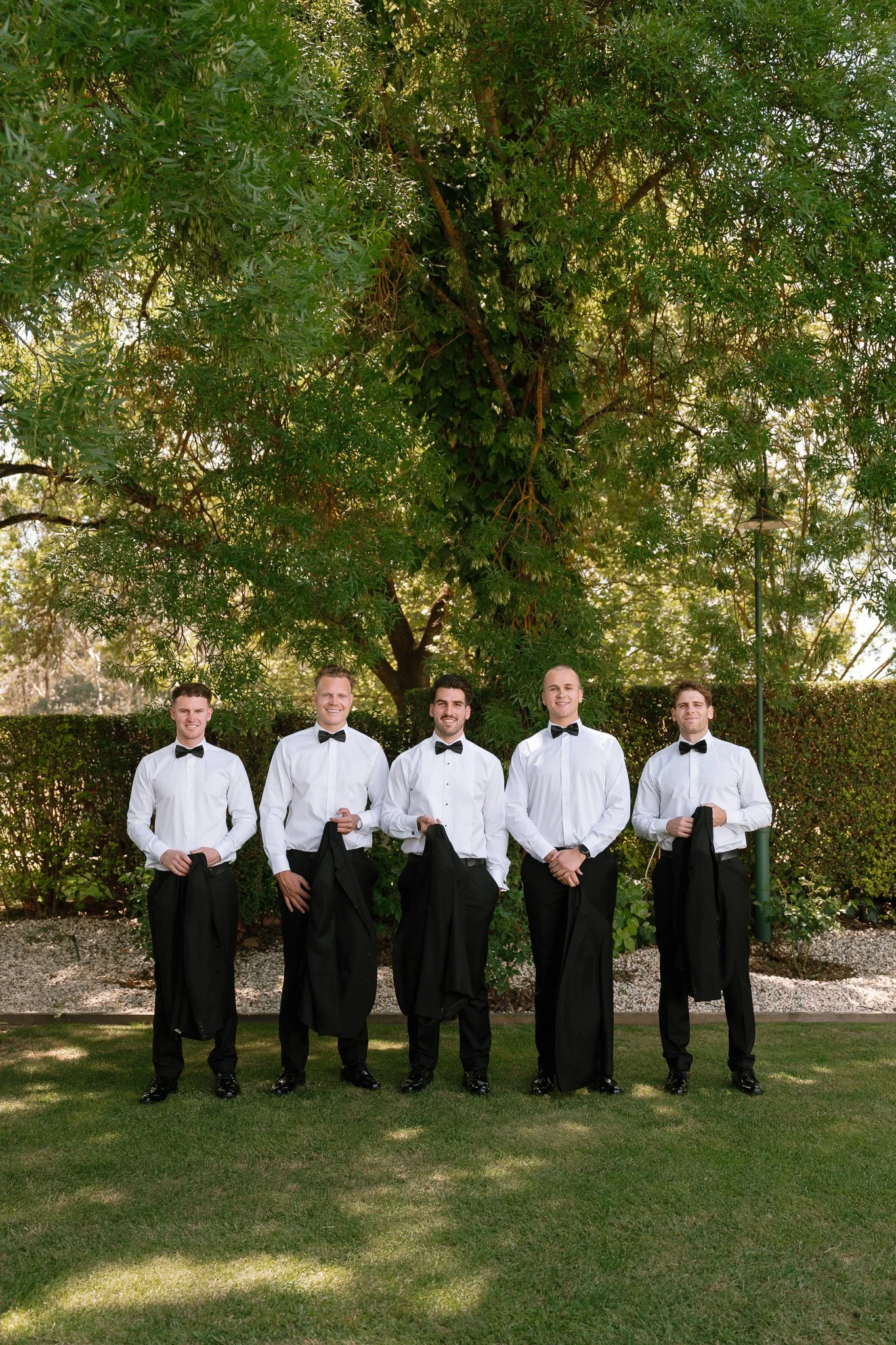 Five men in black tuxedos and bow ties standing outdoors in front of trees and bushes, smiling and holding black jackets.