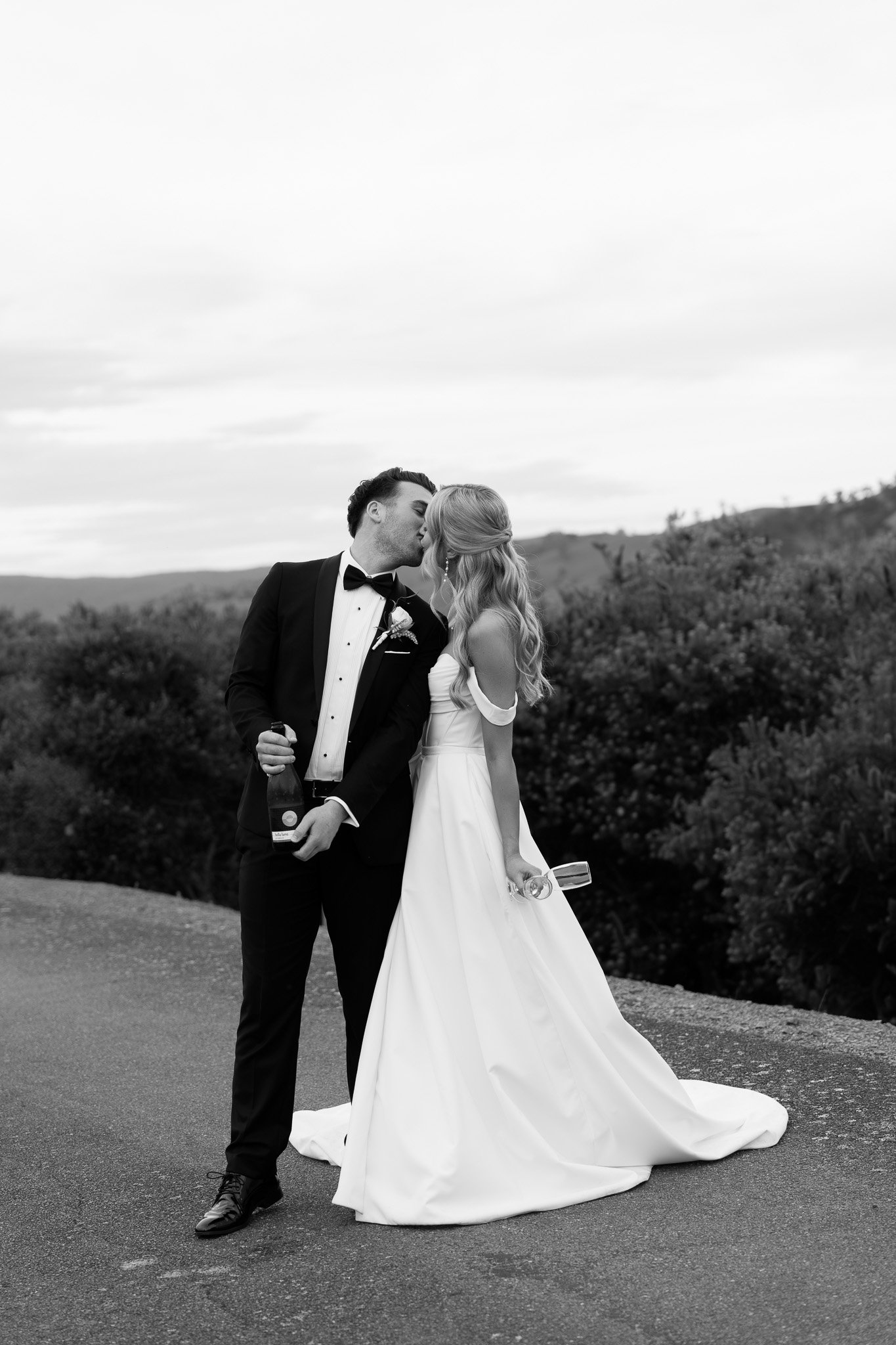 A black and white photo of a newlywed couple kissing outdoors, with the groom in a tuxedo and the bride in a wedding dress holding a glass, standing on a paved road with a natural landscape in the background.