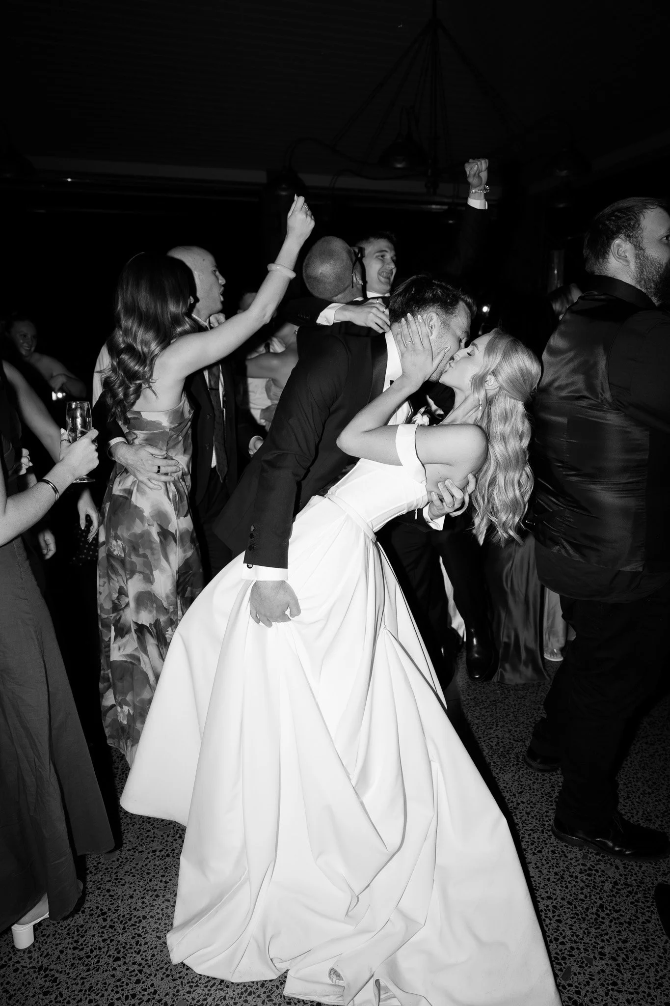 A newlywed couple sharing a kiss at their wedding reception, surrounded by guests celebrating.