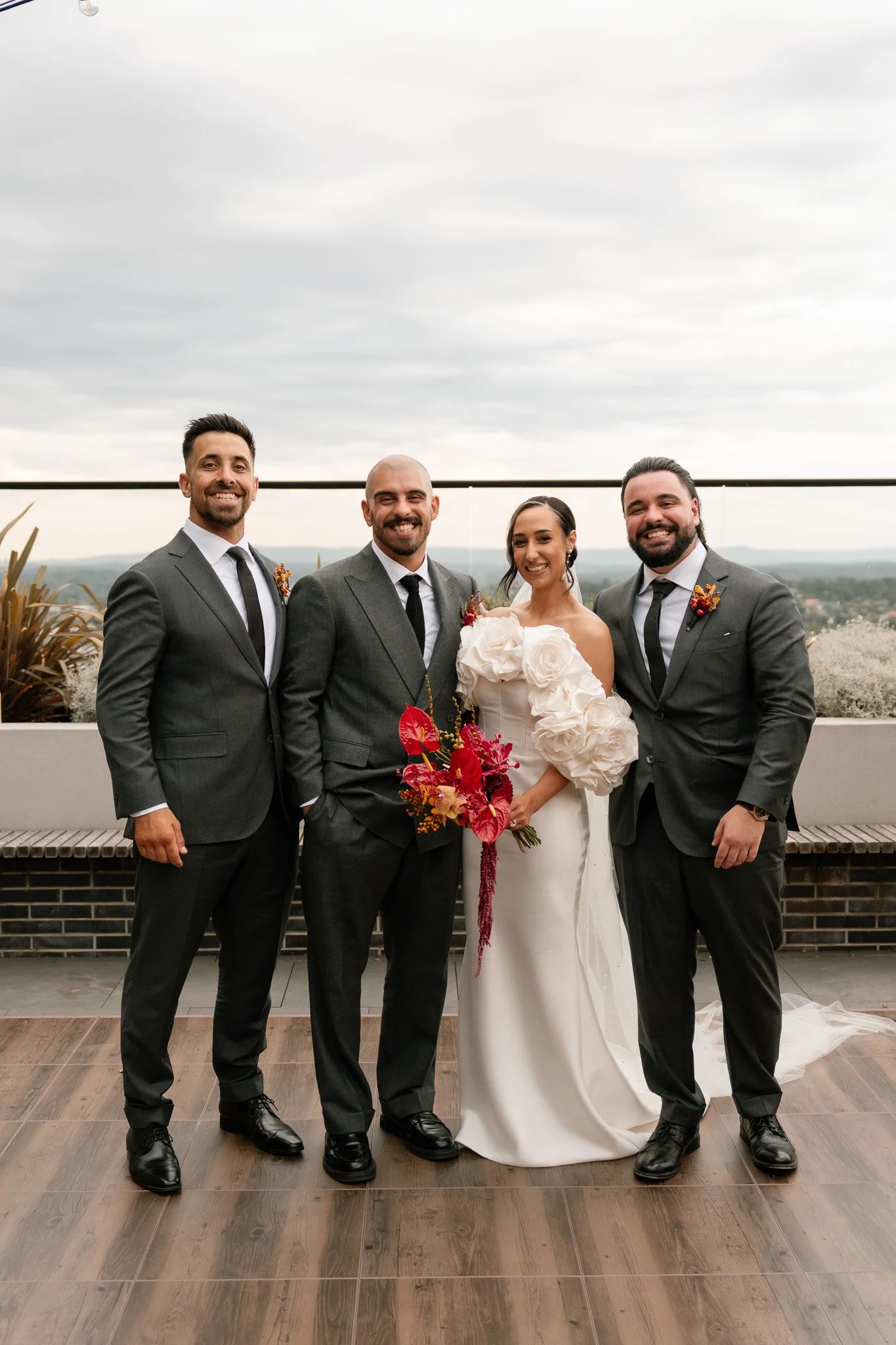A group of four people dressed in formal attire standing on a rooftop with a city view in the background. They are smiling, with the woman holding a vibrant bouquet of red and pink flowers.