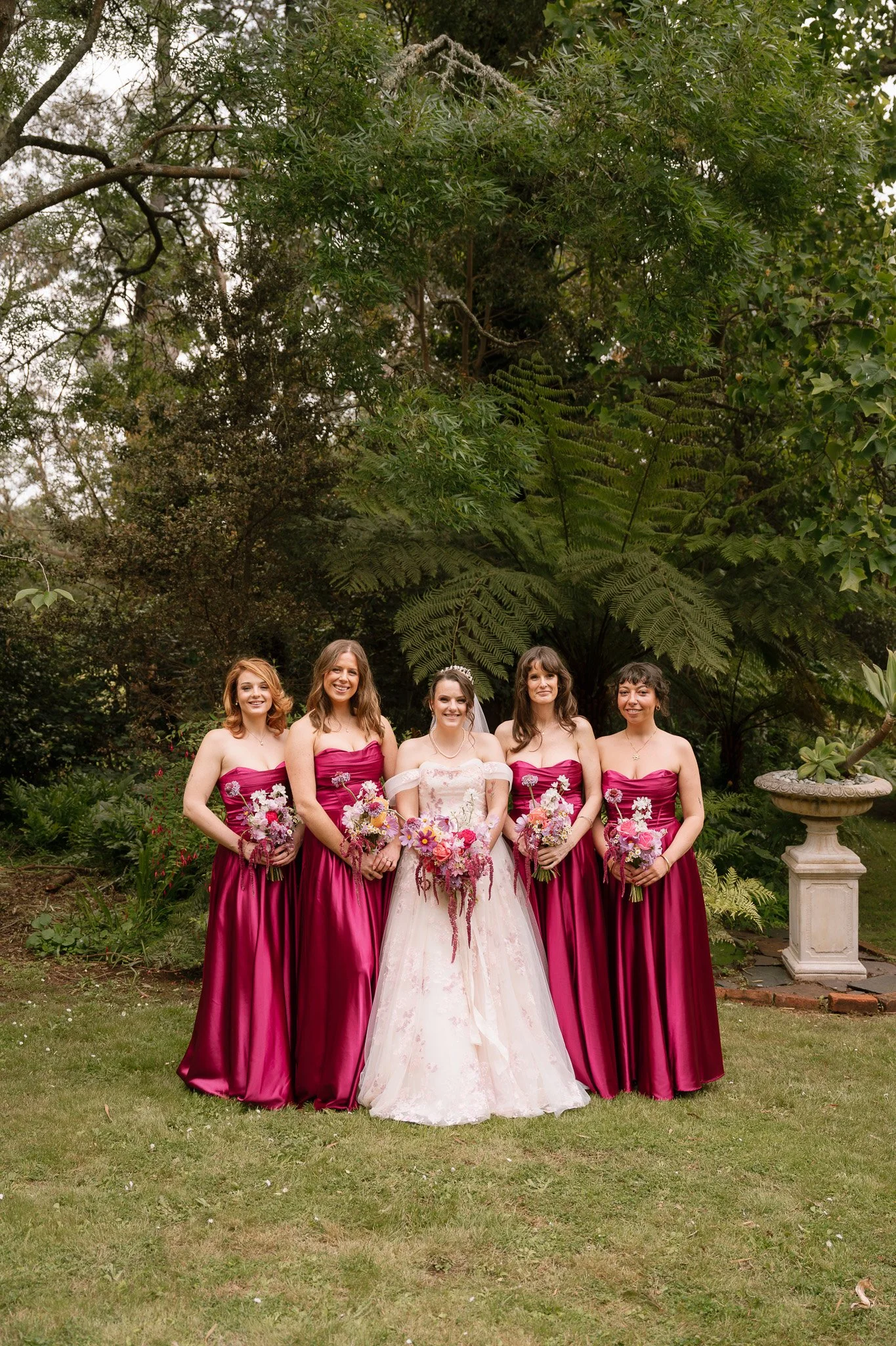 A bride in a white wedding dress with an off-the-shoulder design standing with four bridesmaids in matching deep pink strapless gowns, all holding bouquets of pink, purple, and white flowers, in a garden setting with greenery and a stone urn in the b