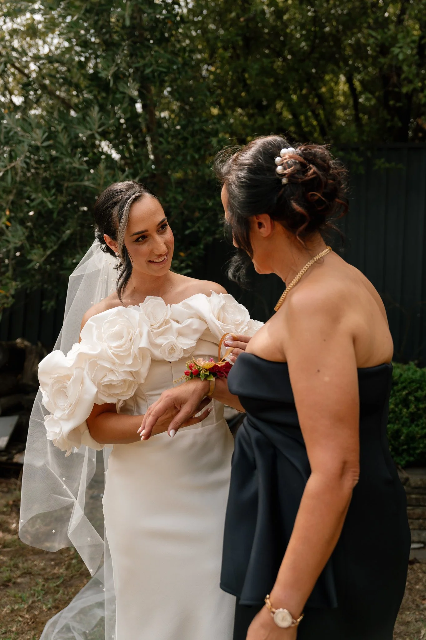 A bride in a white wedding dress with large floral details on the sleeves and a veil, smiling as she receives a corsage from a woman in a black strapless dress in an outdoor garden setting.