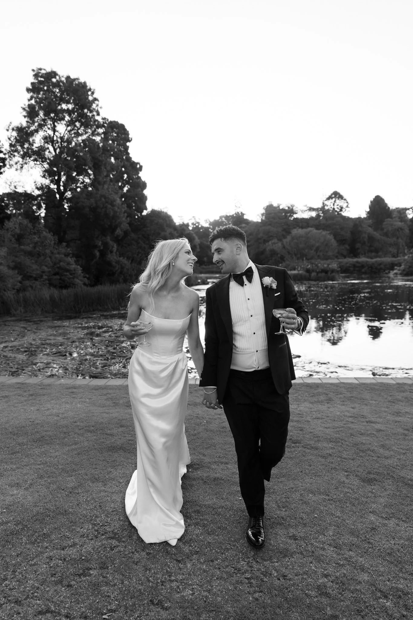 A newlywed couple in wedding attire walking hand in hand outdoors, smiling at each other near a lake with trees in the background, in black and white.