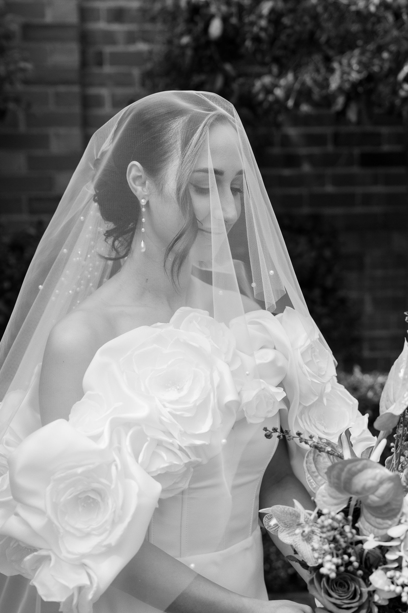 A bride wearing a veil and earrings, holding a bouquet of flowers, smiling softly with her eyes closed outdoors.