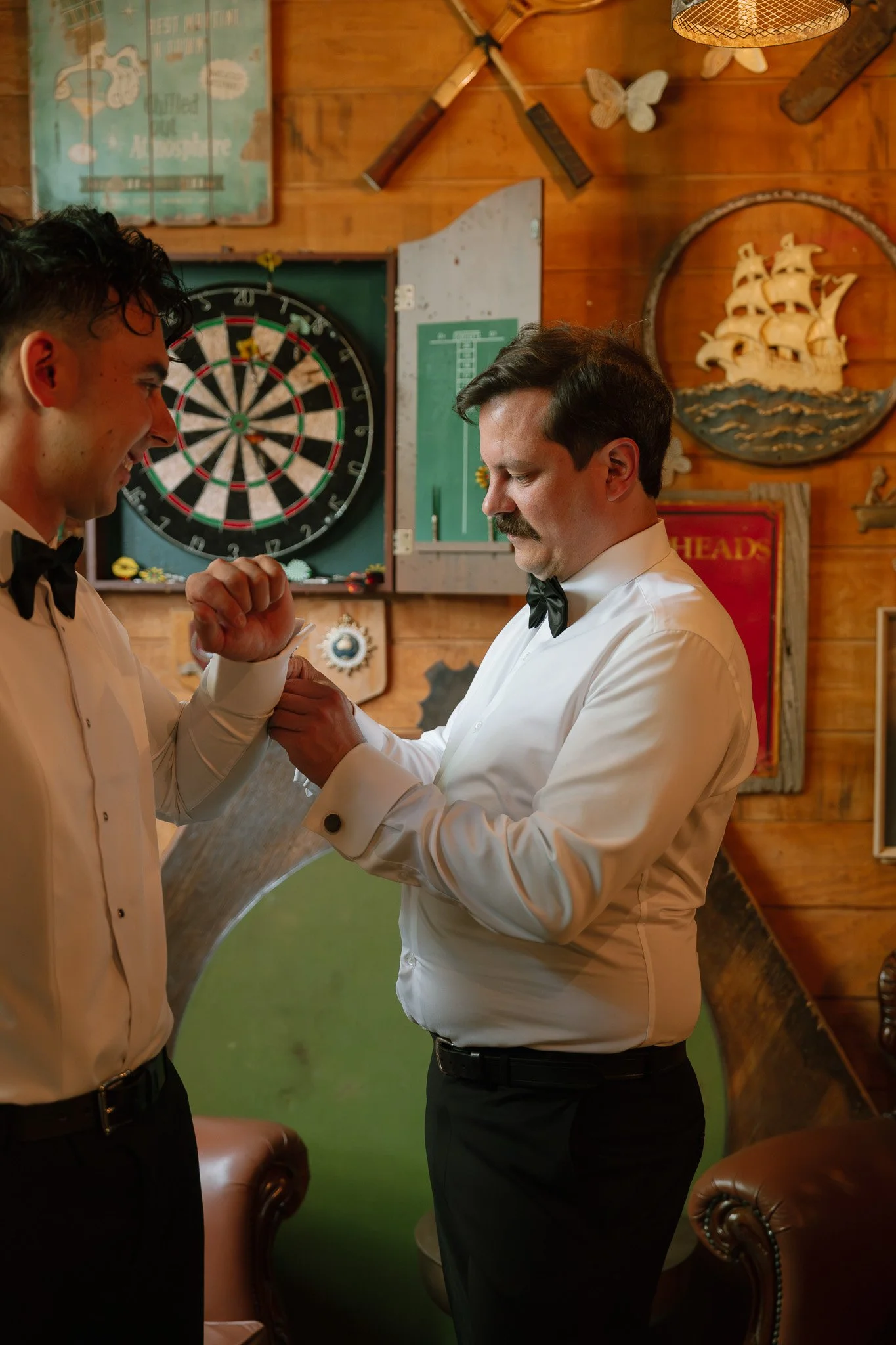 Two men in tuxedos with bow ties, one younger and one older, in a cozy, wood-paneled room decorated with nautical and vintage signs. They appear to be exchanging a wristband or cufflink.