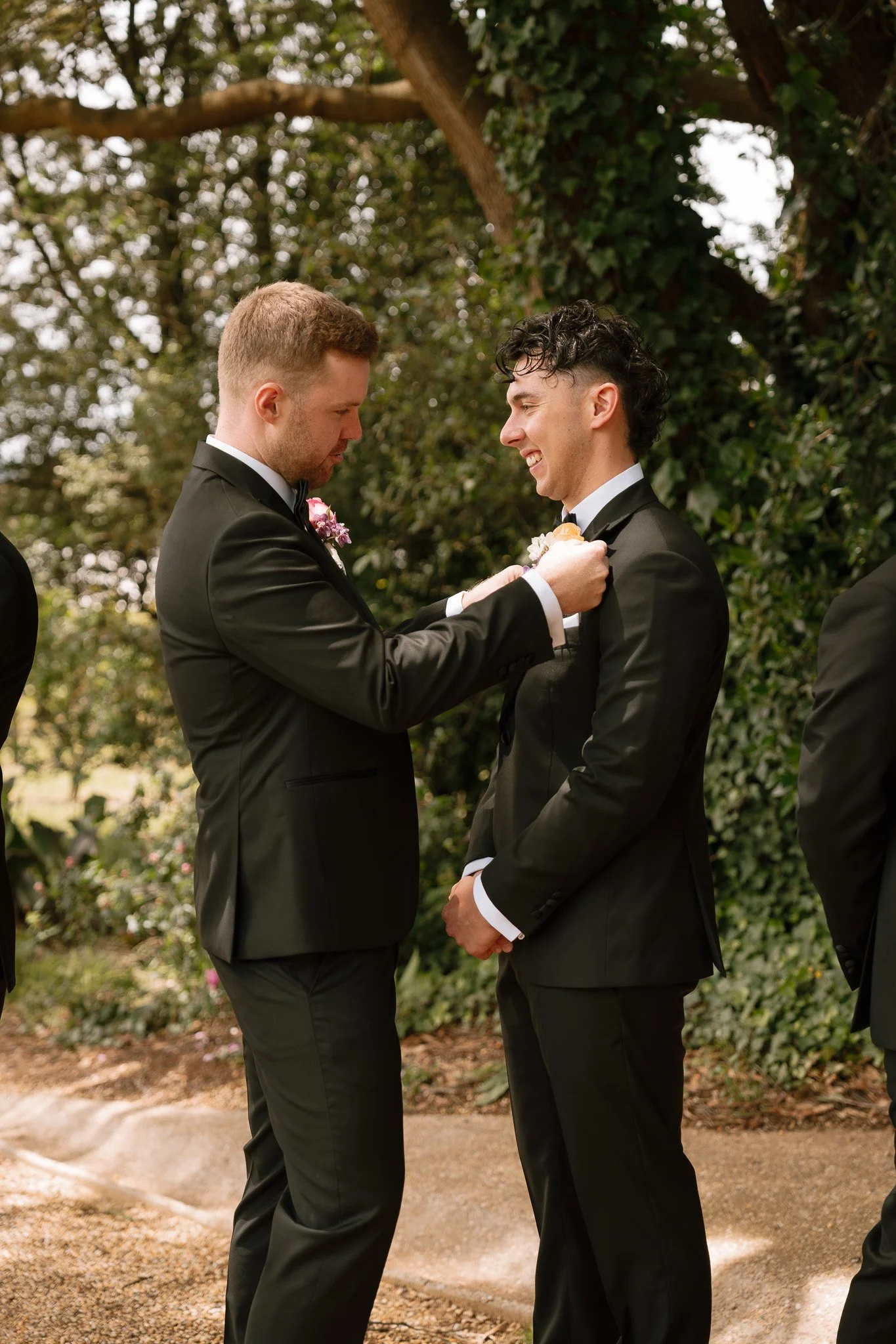 Two men in black tuxedos at a wedding, with one man pinning a boutonniere on the other, outdoors with trees and greenery in the background.