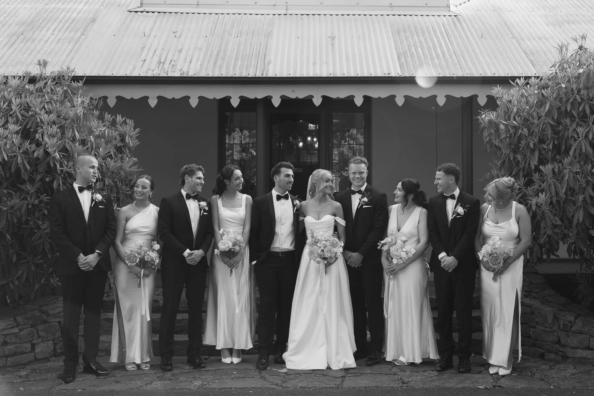 Black and white photo of a wedding party with bride, groom, and six bridesmaids and groomsmen standing outdoors in front of a building with trees and stone pathway.