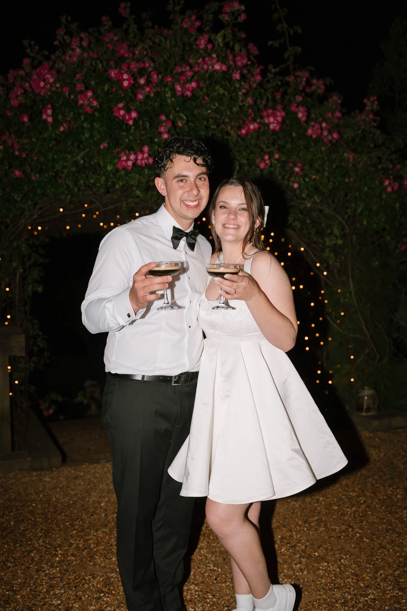 A young man and woman in wedding attire celebrating at night, holding glasses of champagne, standing under an arch decorated with pink flowers and string lights.