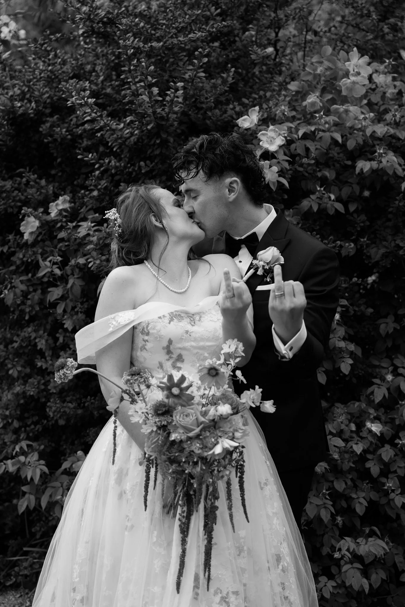 A black and white photo of a bride and groom kissing outdoors, the bride holding a bouquet of flowers, both making a gesture with their hands.
