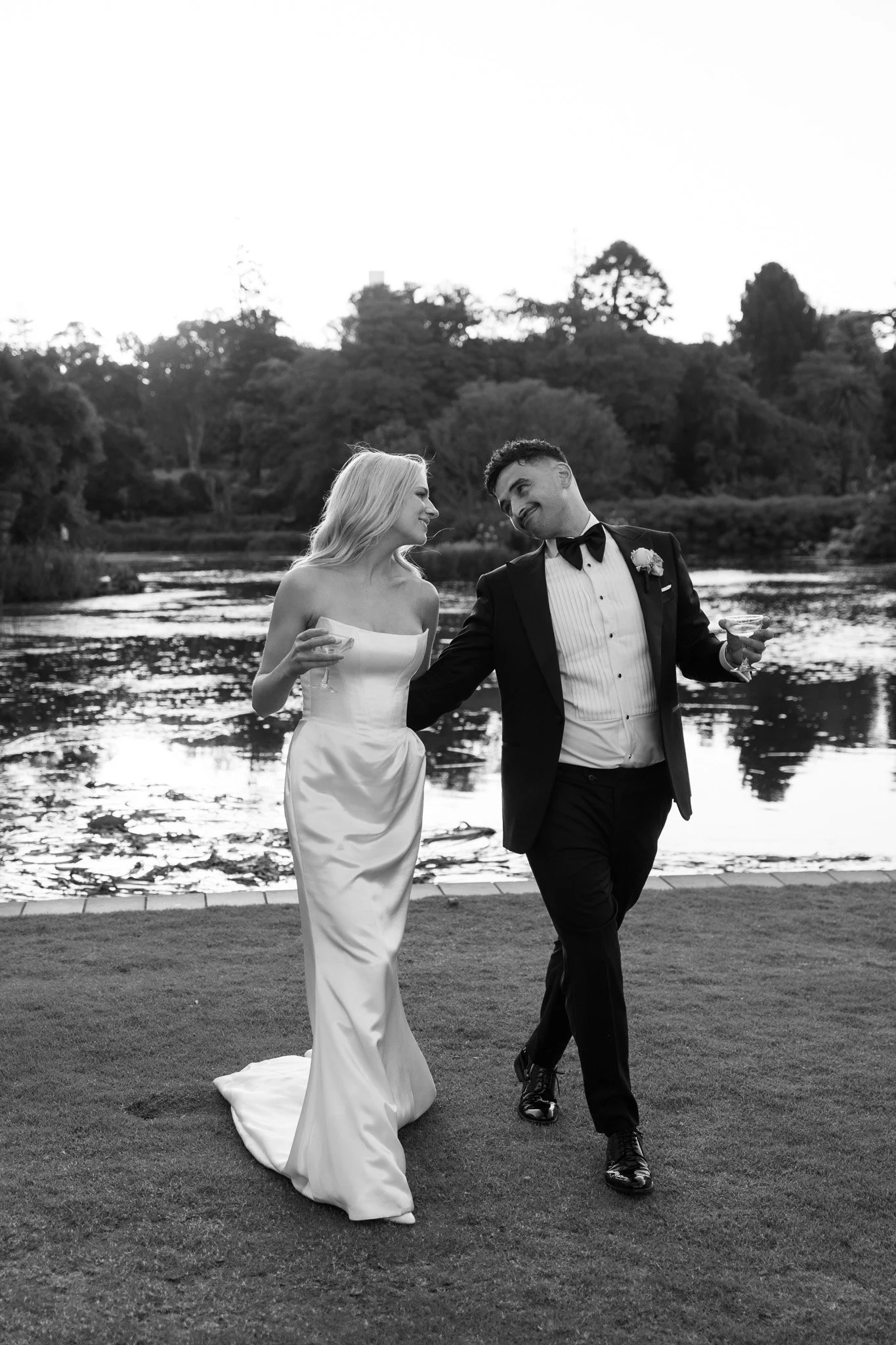A black and white photo of a bride and groom walking by a river, holding drinks, smiling, and looking at each other, outdoors with trees in the background.