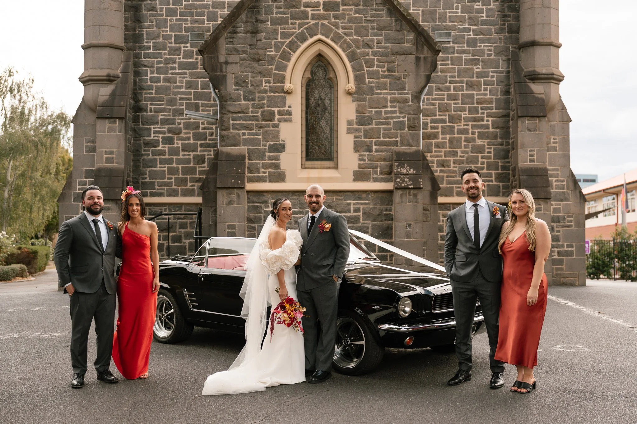 Group of six people, including bride and groom, posing in front of a vintage black convertible car outside a stone church.